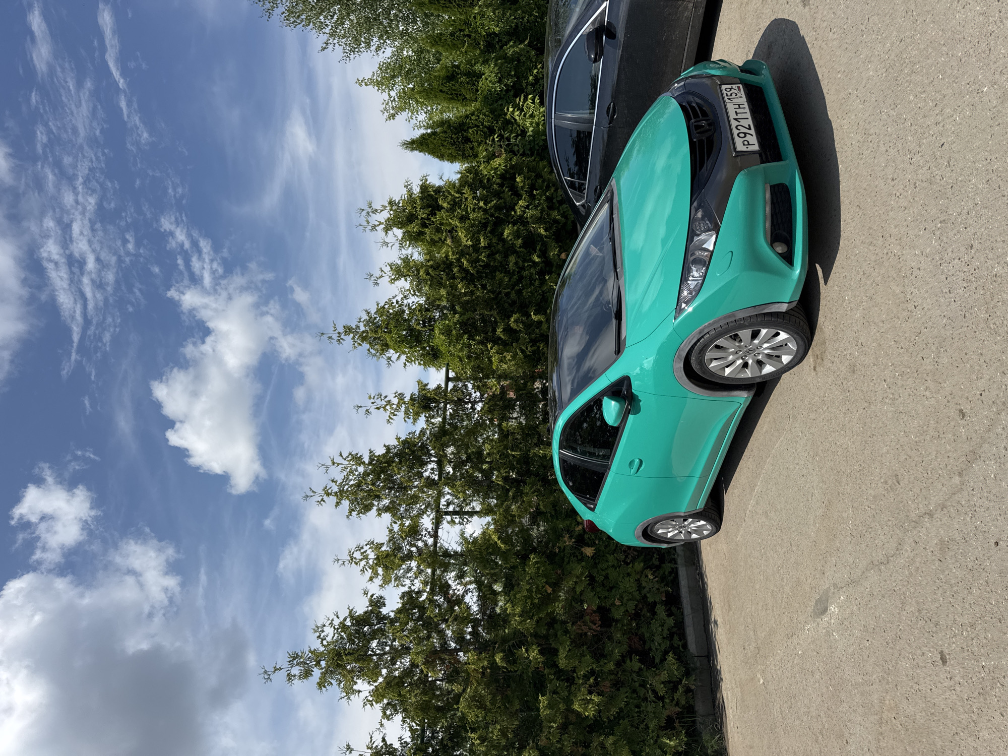 A turquoise Honda Civic is parked on an asphalt surface in front of a row of evergreen trees. The car has a black grille and a license plate that reads "P921TH 159". A black sedan is parked next to it. The sky is blue with scattered white clouds. The scene is likely a parking lot in Chaikovsky, Russia.