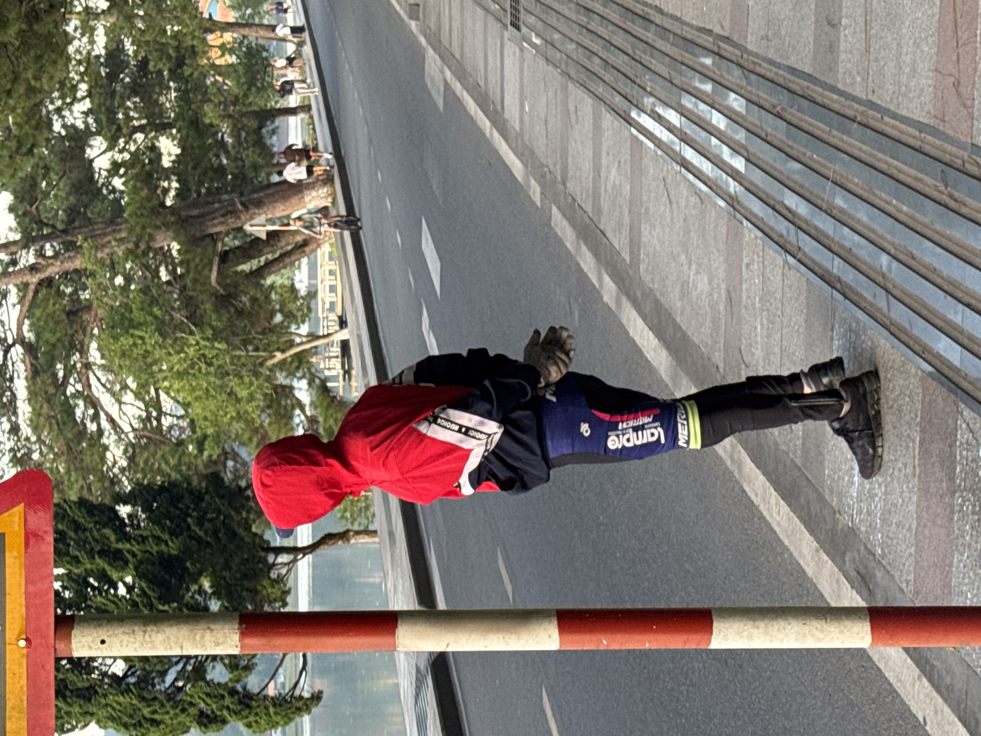 In Da Lat, Vietnam, a person stands by a red and white striped pole next to a street. The person is wearing a red hooded jacket, black pants, and black athletic shoes. They are also wearing gloves. The street is empty except for a few distant figures walking and a cyclist. The background shows a body of water and pine trees. The pavement has tactile paving.