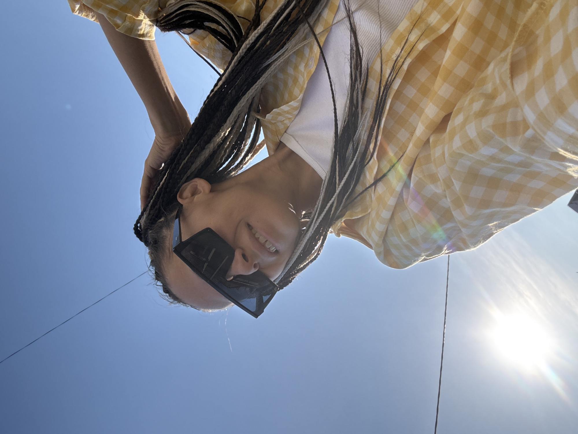 A low-angle photograph depicts a person in Fedorino, Russia, wearing black sunglasses, a white tank top, and a yellow gingham shirt. The person has braided hair with a mix of black and white strands and is holding their hair with their hand. The background consists of a clear blue sky with the sun visible in the lower right corner. A thin, dark line, possibly a power line, is visible in the sky.