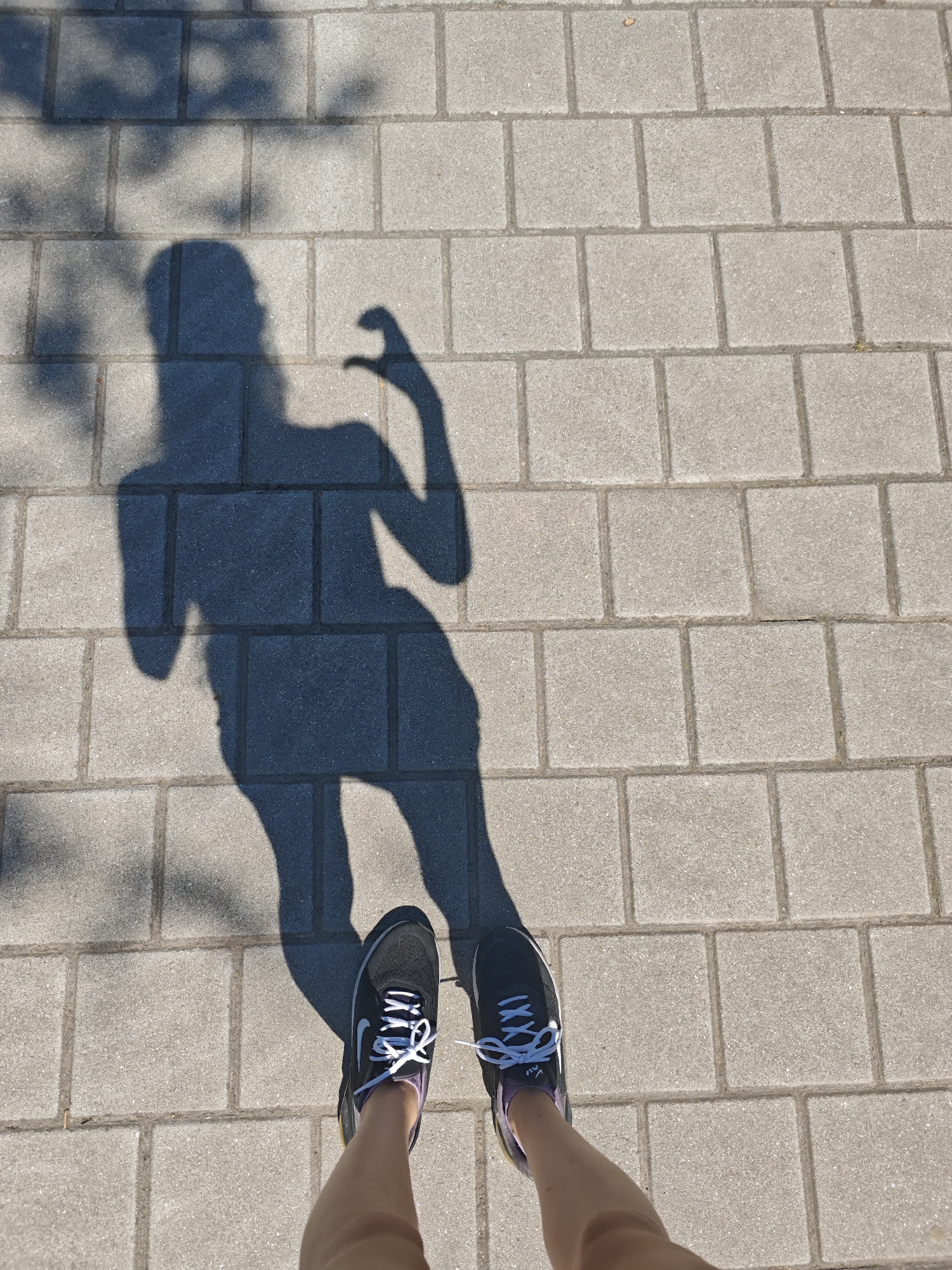 An overhead shot shows a shadow of a person in athletic shoes, standing on a paved area. The shadow is of a person with their right arm raised and their hand in a heart shape. The shoes appear to be Nike brand. The paving stones are rectangular and laid in a repeating pattern. The scene suggests an outdoor environment, possibly a park or walkway, with dappled sunlight casting shadows from trees onto the paving. The location is specified as Tilburg, Netherlands.
