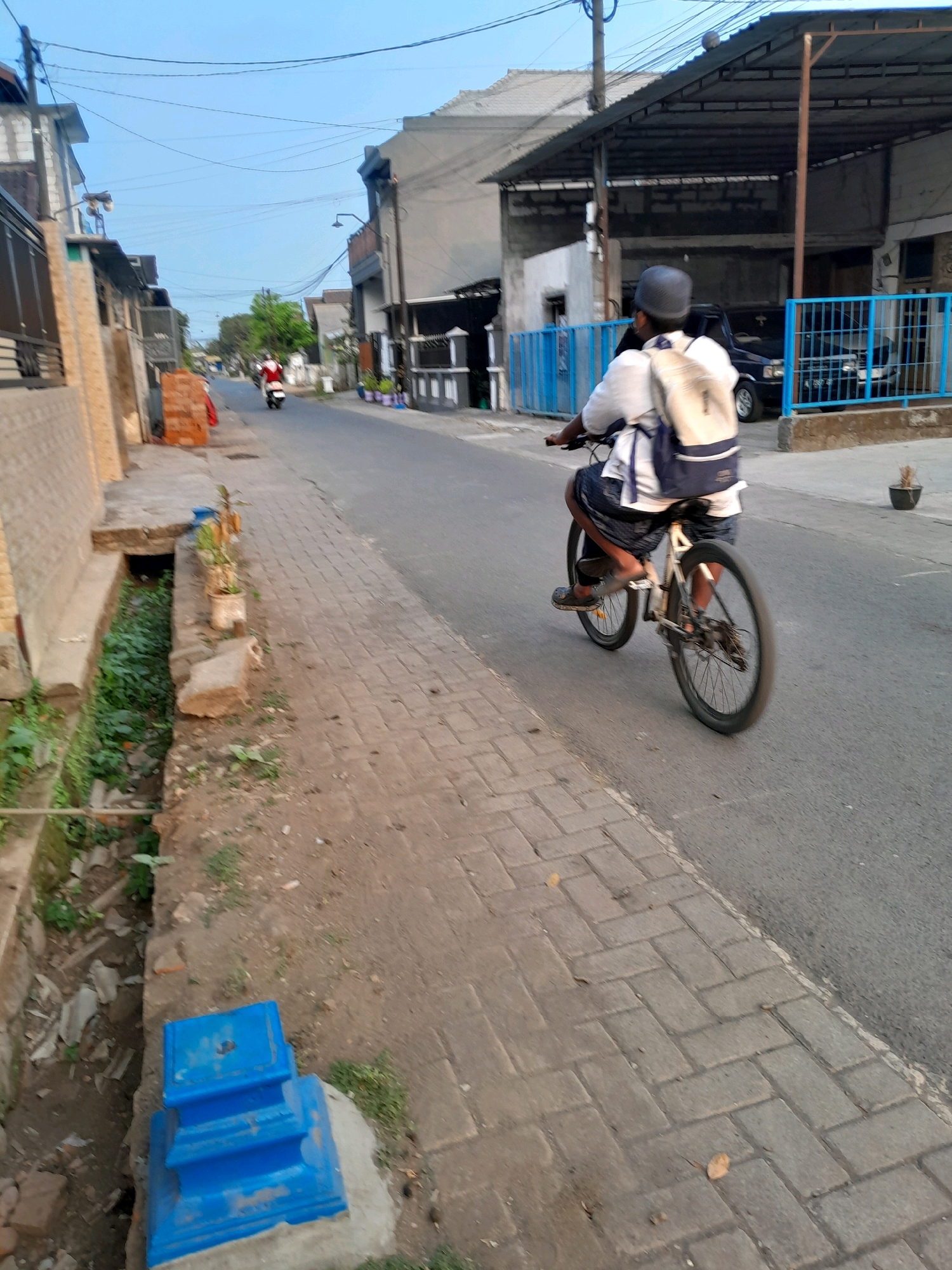 The image depicts a street scene in Ngadilangkung, Indonesia. In the foreground, a person wearing a white shirt, dark shorts, and a backpack is riding a bicycle on the asphalt road. The person is also wearing a dark helmet. To the left of the road, a paved sidewalk runs alongside a narrow, overgrown drainage ditch. Various structures, including houses and a covered carport with a blue metal fence, line the street. In the background, another person is seen riding a motorbike on the road. The sky is a clear blue, suggesting it is daytime. The overall environment appears to be a residential area within the city.