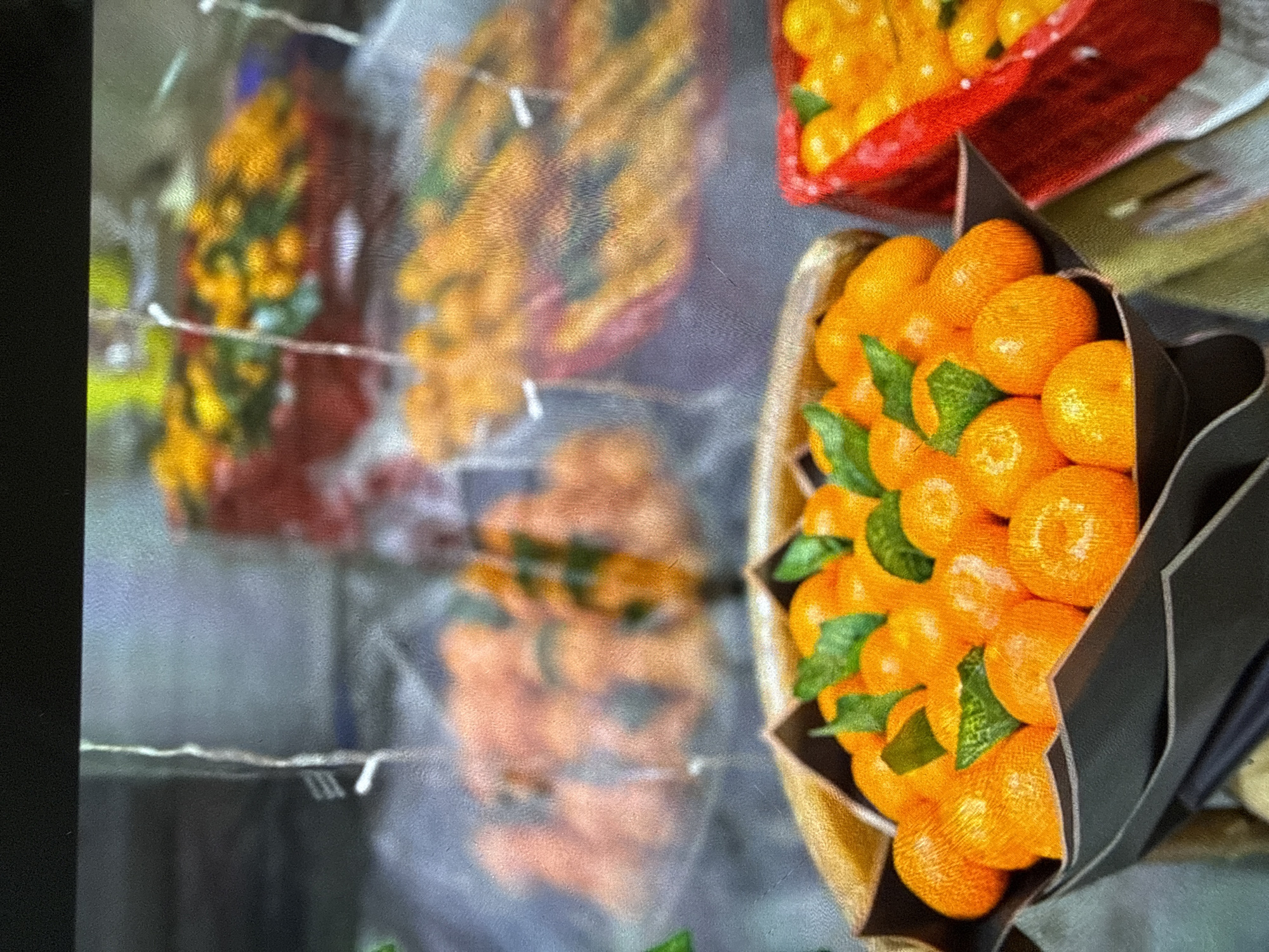 A bouquet of tangerines is presented in a dark wrapping with green leaves interspersed among the fruit. In the background, out of focus, are red baskets filled with tangerines and other orange-colored fruits. The lighting suggests an indoor setting, possibly a market or storefront. The image is of low resolution and appears to be a photograph or a digitally captured image. The scene takes place in an unknown location within an unknown city.