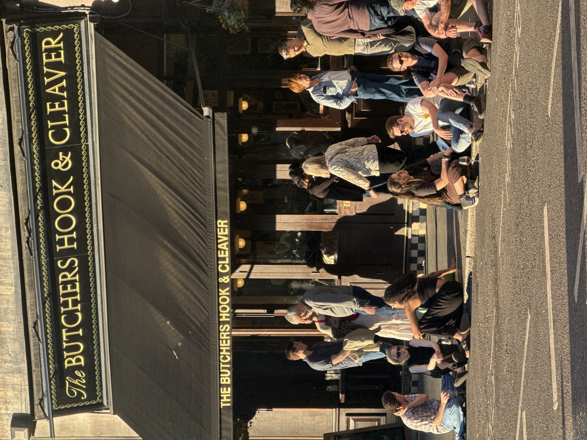 A horizontal eye-level medium shot depicts a group of people outside "The Butcher's Hook & Cleaver," a pub in London, United Kingdom. The pub's awning and signage are prominent on the left. A row of people sits outside the pub in the street. The street is paved and has painted road markings. The scene suggests a casual outdoor social gathering.