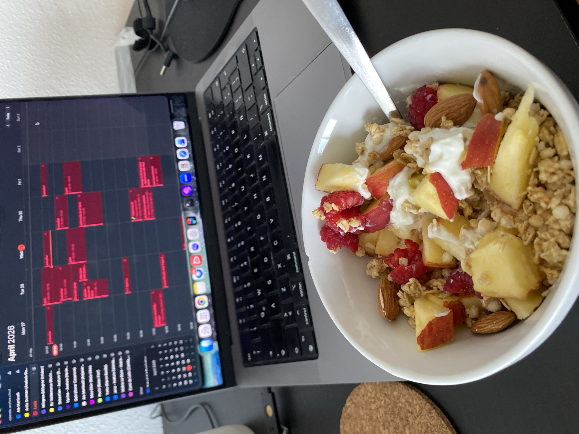 The image presents an overhead view of a desk setup, featuring a breakfast bowl next to an open laptop. A white bowl on the right is filled with a nutritious meal consisting of yogurt, granola, diced red apples, diced yellow fruit (possibly pineapple), vibrant red raspberries, and whole almonds. A silver spoon is resting inside the bowl.
To the left, a silver MacBook Pro laptop is open, its screen displaying a calendar application. The visible calendar shows "April 2026" with a weekly view from "Mon 27" to "Sun 3". Numerous red blocks populate the calendar grid, indicating scheduled events or busy periods. A sidebar on the left lists various calendar entries such as "calendar avature cloud", "Ale Martinelli", "Anita Guzman", "Lucia", "Milagros (Milagros Dziuban)", "Ro Valdemores", "Sebastian - Avature", "Vanina - Avature", "Oili (Oscar Casanova)", "Ale Martinelli + Alejand...", and "Pachi (Belen Fiel)". A vertical dock or toolbar on the right side of the screen displays application icons, including Mail, Calendar, Safari, Messages, Google Chrome, and others. The laptop's keyboard is visible, with keys like "option" and "command" clearly distinguishable.
The items are situated on a dark-colored desk surface. A circular cork coaster is partially visible beneath the bowl. Wires and cables can be seen in the background, near the laptop's hinge. The scene suggests a morning routine of having breakfast while engaging in work or planning activities, likely within an indoor setting in Gijón/xixón, Spain. The detailed calendar for April 2026 implies a focus on future scheduling.