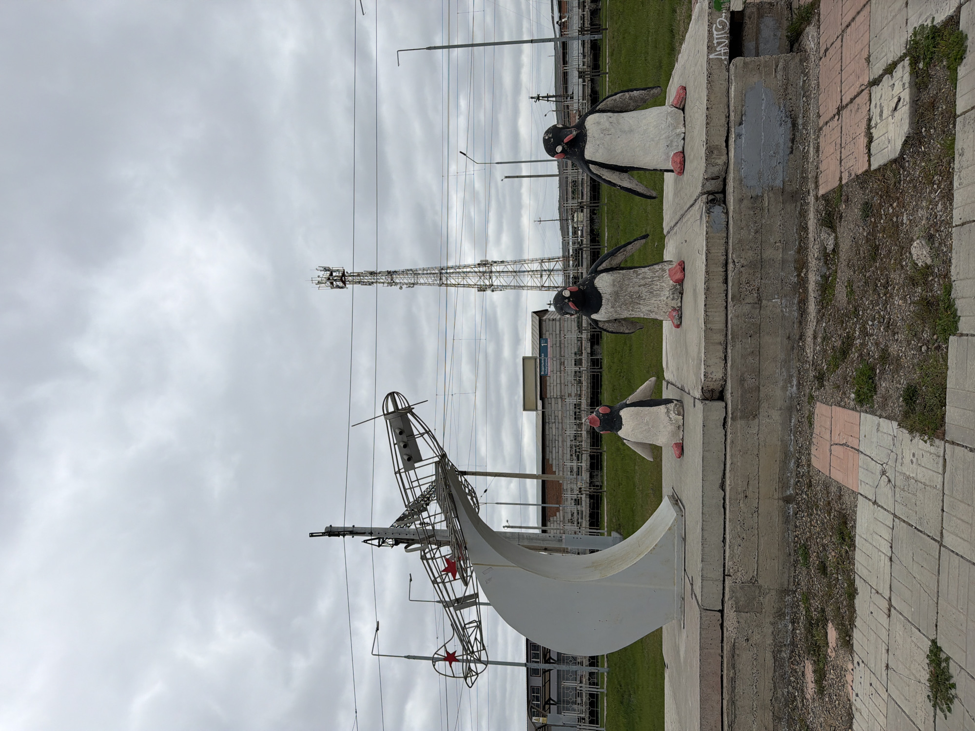 The image shows an outdoor scene in Krasnoyarsk, Russia. Three penguin statues stand on concrete blocks near a paved area with grass visible in the background. Behind the penguins is a metal sculpture resembling an airplane or ship framework, with red star details. A tall transmission tower is visible in the background, along with buildings and utility poles. The sky is overcast with grey clouds.