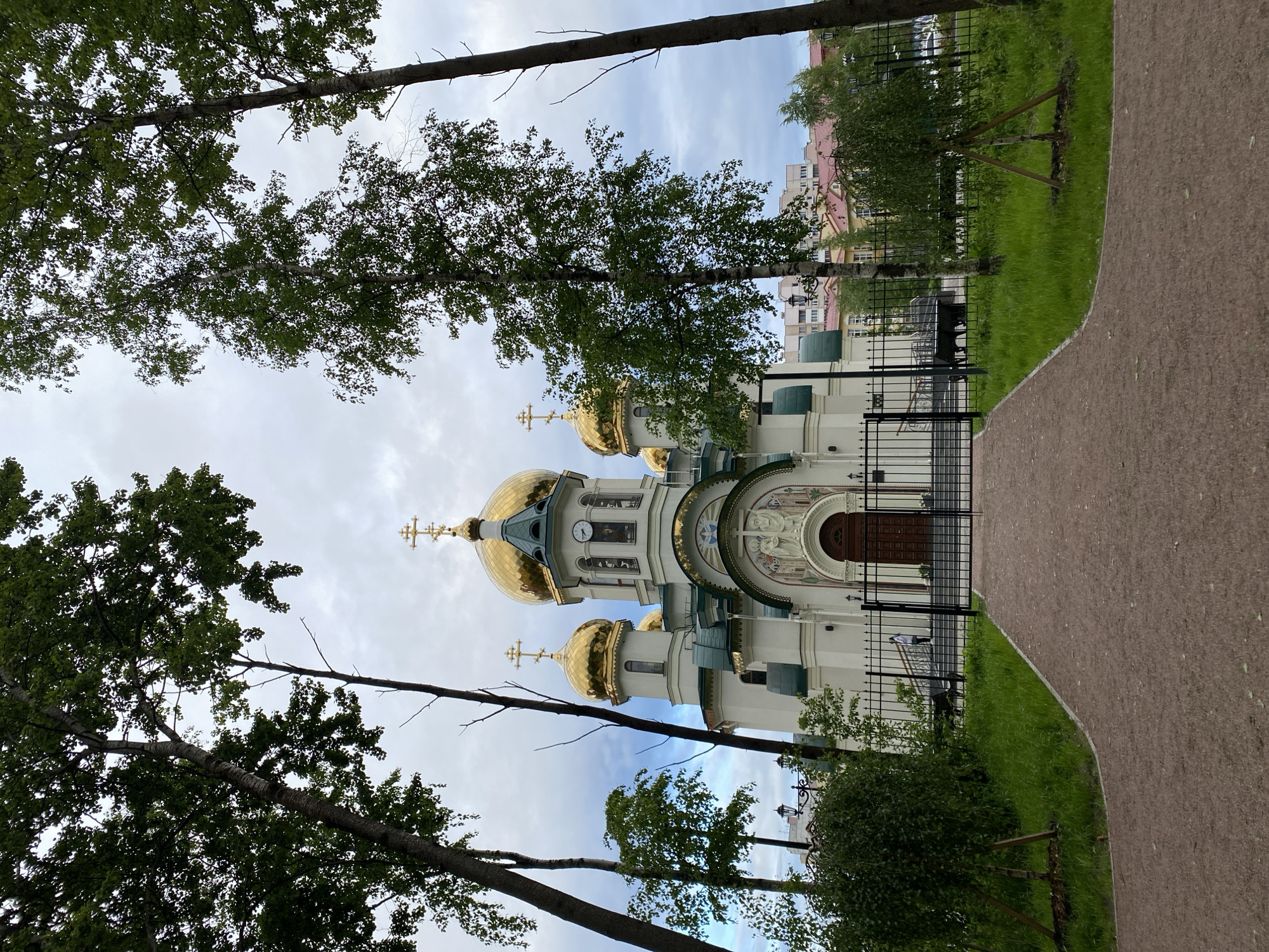 The image captures a view of the Church of the Holy Martyr Valeria in the city of Saint Petersburg, Russia. The church features white walls, multiple golden domes, and crosses. The architectural style exhibits classical Russian Orthodox features. The building is enclosed by a black metal fence and is adjacent to a paved pathway. Trees are partially obscuring the view of the church, with the sky visible through the branches. Buildings are visible in the background.