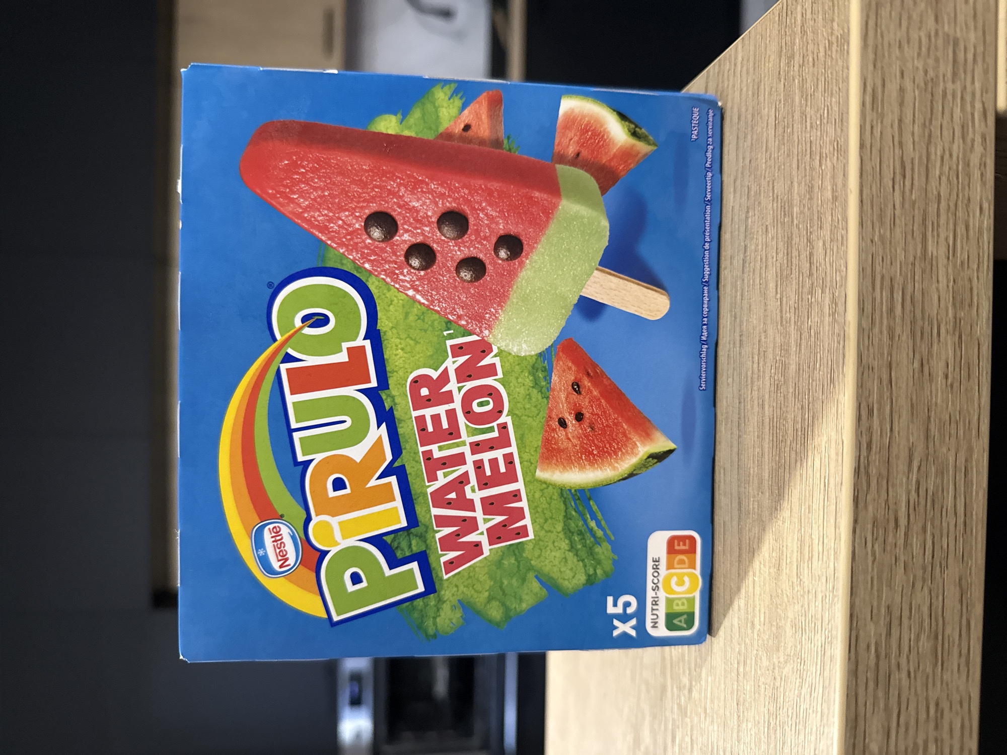 A rectangular Nestlé Pirulo Water Melon popsicle box is centrally positioned on a light brown, laminated wooden surface. The box features a bright blue background with a prominent multi-colored "Pirulo" logo. Below the logo, "WATER MELON" is printed in red lettering with small black seed illustrations. A large graphic of a red and green watermelon-shaped popsicle, complete with a wooden stick and five dark, circular 'seeds', is depicted on the front. Two smaller watermelon slices are partially visible in the background of the box design. The bottom left corner displays "x5" and a "NUTRI-SCORE C" label, indicating the product's nutritional rating. The background behind the box is dark and out of focus, revealing a domestic interior, possibly a kitchen, with vague shapes of appliances visible. The lighting is artificial, illuminating the box and the immediate foreground. The scene is static, captured in the city of Unknown, Unknown.
