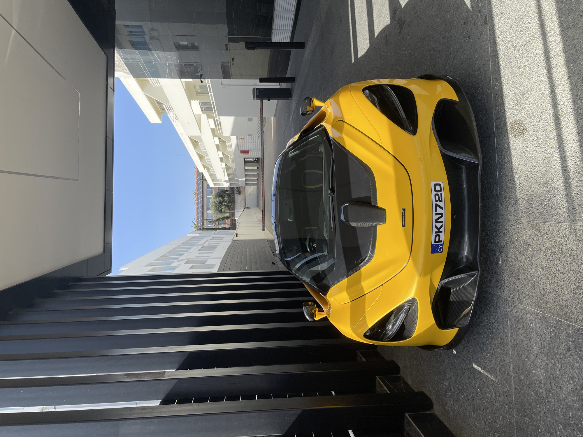 A yellow McLaren 720S is parked in what appears to be the entrance of a building in Ayia Napa, Cyprus. The vehicle's front is visible and has a license plate with "PKN720" displayed. The building surrounding the car has a modern design with horizontal black bars. The sky is clear and blue.