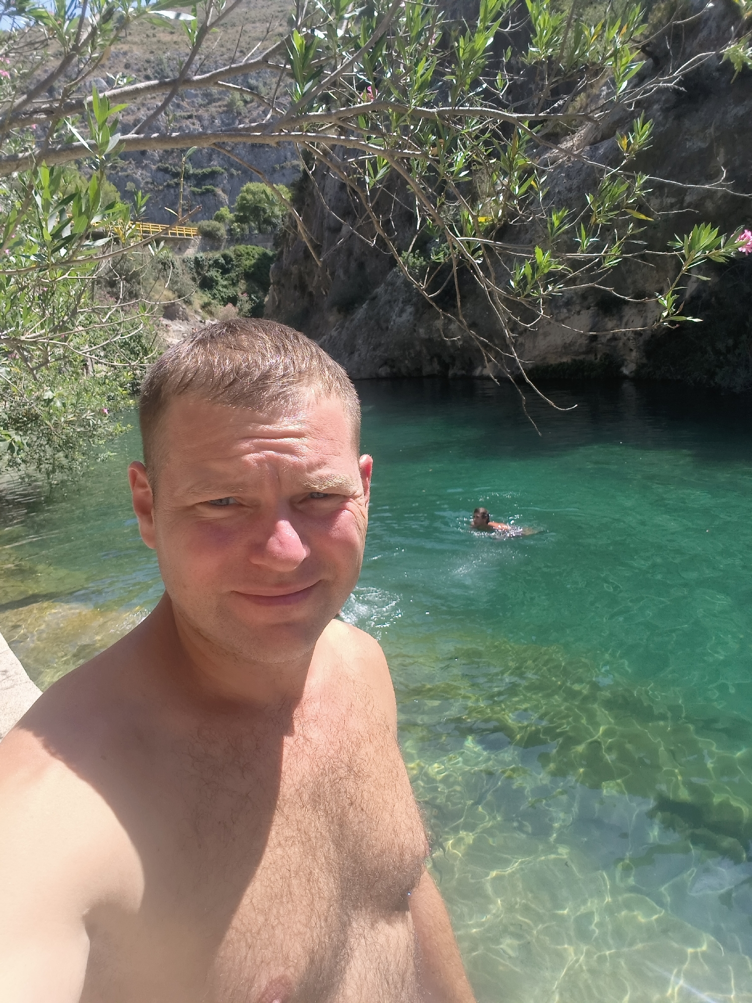 In this selfie, a shirtless Caucasian man with short blond hair and blue eyes is positioned in the foreground, looking towards the camera with a slight smile. He appears to be in a body of water, with his upper torso and face visible. Behind him, the water is a clear, vibrant turquoise, revealing a sandy or rocky bottom. In the middle ground, another person, appearing as a silhouette from this distance, is swimming in the water. The background features a lush green environment with trees and rocky cliffs. A yellow structure, possibly a bridge or walkway, is visible on the cliffside in the distance. The lighting suggests a sunny day, with the sun casting shadows on the man's chest. This scene is likely the Pou Clar riverbed in Ontinyent, Spain, known for its natural pools and picturesque surroundings.