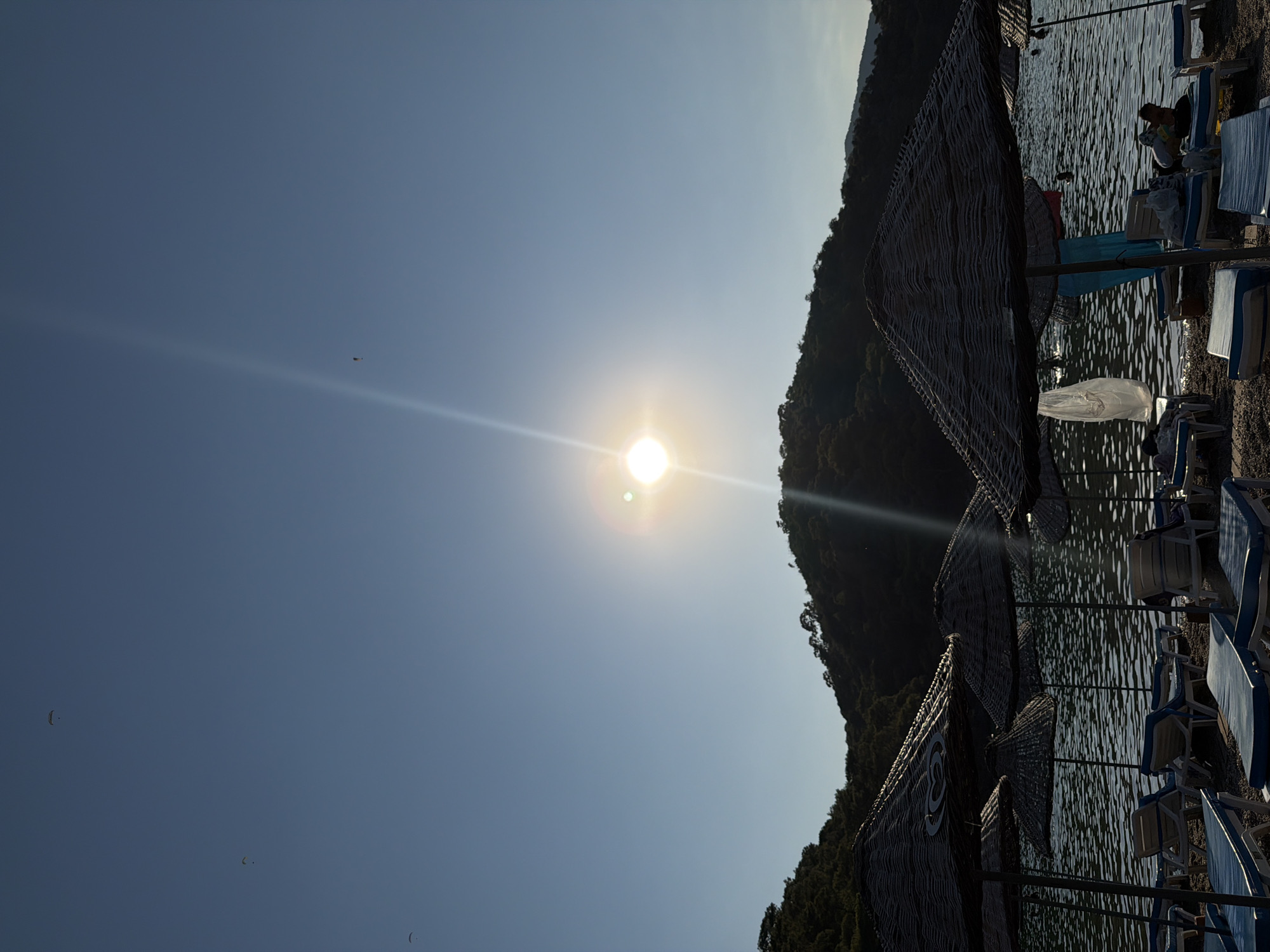 This image depicts a beach scene at sea, likely in Fethiye, Turkey, given the characteristic Mediterranean landscape with lush green hills meeting the water. The sun is prominently positioned in the upper central part of the frame, casting a bright glare and a sunbeam that extends downwards. In the distance, a silhouette of a tree-covered hill is visible against the clear blue sky. In the foreground, several beach umbrellas made of natural straw material are erected over empty lounge chairs arranged on the pebbly shore. The sea, visible behind the umbrellas, is calm with gentle ripples. The water appears dark due to the backlight from the sun. There are a few people visible in the water, appearing as small dark shapes, possibly swimming or wading. A white plastic bag hangs from one of the umbrellas. The overall atmosphere suggests a late afternoon or early evening scene on a sunny day at a coastal resort. Three paragliders are visible in the sky, indicating possible recreational activities in the area.