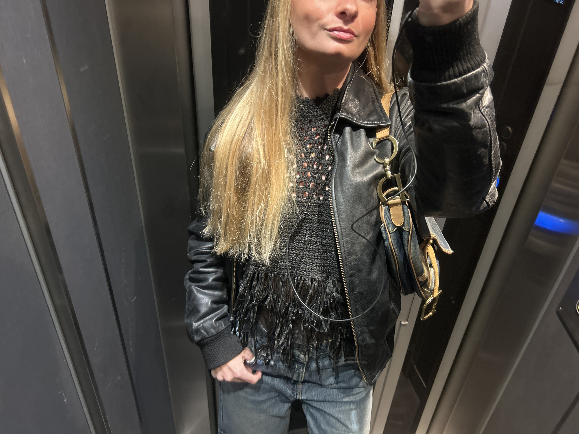 A person stands inside an elevator. The person wears a black leather jacket over a black, fringed top and blue jeans. A denim handbag with brown leather trim hangs from their shoulder. They appear to be holding a device in their left hand, possibly attached to earphones which are visible around their neck. The elevator walls are gray with vertical lines and a blue light is visible on the right wall. The location appears to be indoors, possibly in a building within the city of Unknown, Unknown.