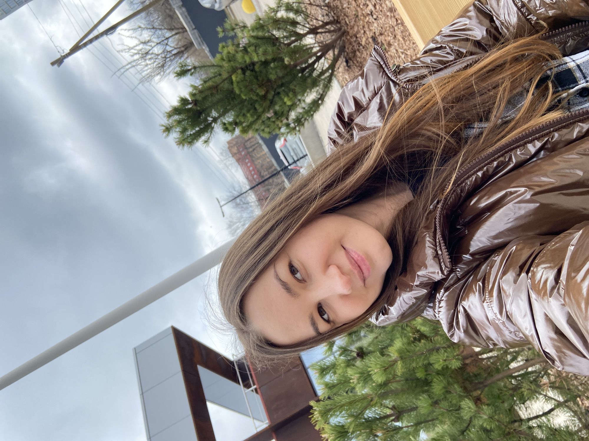 A young woman is featured in an outdoor selfie, likely taken in Izhevsk, Russia. The image is composed at an angle, capturing her head and upper torso. She has long, light brown hair and gazes directly at the camera with a gentle, friendly smile. She is dressed in a shiny, dark brown or bronze puffer jacket, worn over a black and white plaid shirt, visible at the collar and sleeve.
The background presents a mix of urban and natural elements under a heavily overcast, cloudy sky. Evergreen trees are visible to her right and in the lower left portion of the image. In the mid-ground, a bare-branched tree and utility poles with power lines suggest cooler weather, possibly late autumn or winter. To her left, a modern building with stark white and rust-colored (or similar material) architectural features can be seen. The ground cover consists of brown material, possibly mulch or dried leaves. The overcast conditions indicate daytime with diffused light. No visible text is present in the image.