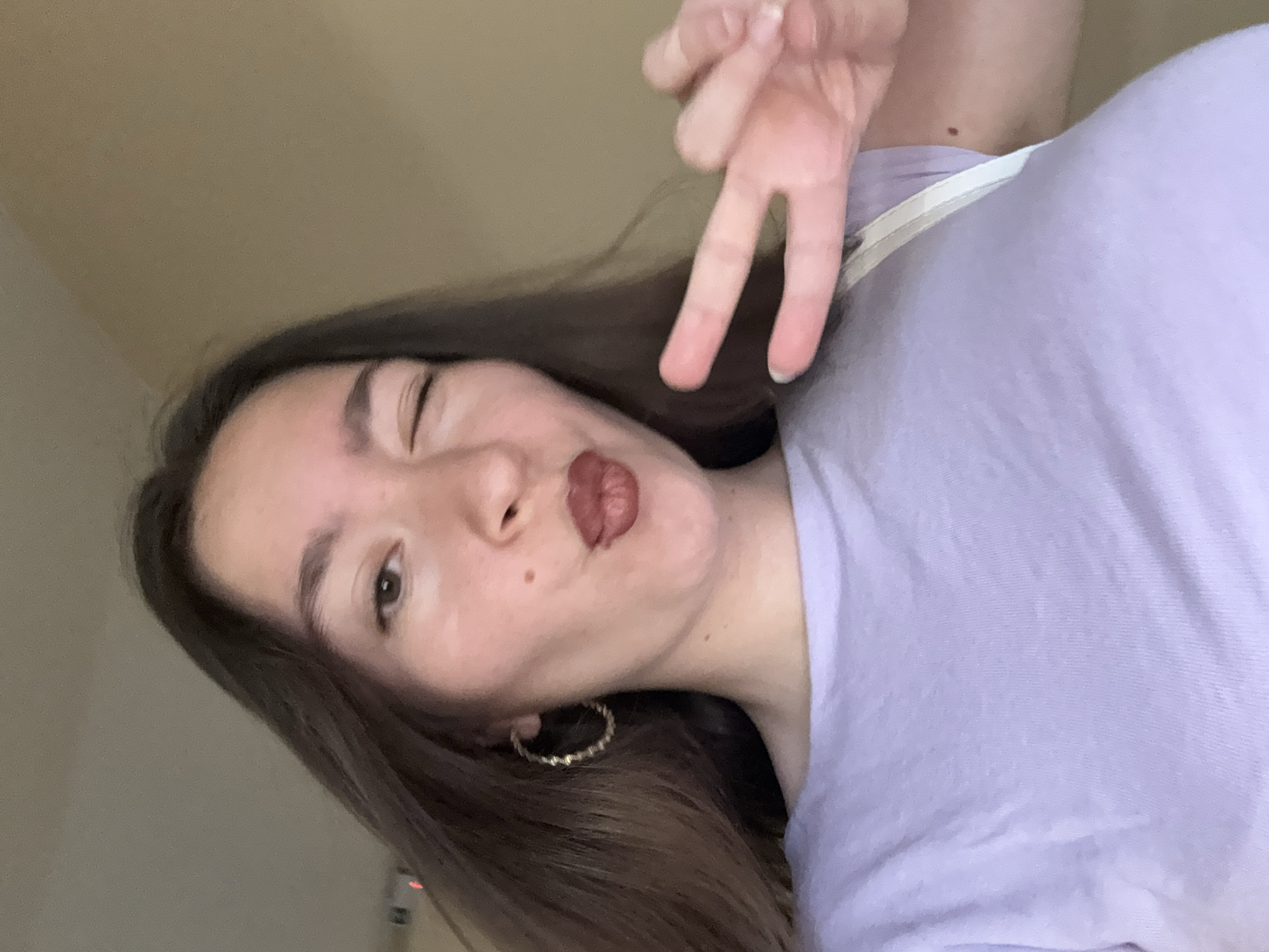 A young woman is taking a selfie. She is wearing a light purple t-shirt and a large gold hoop earring. Her lips are pursed, and she is winking at the camera. Her right hand is held up in a peace sign. The background is a plain, light brown wall. The image appears to have been taken indoors, possibly in a residential setting.