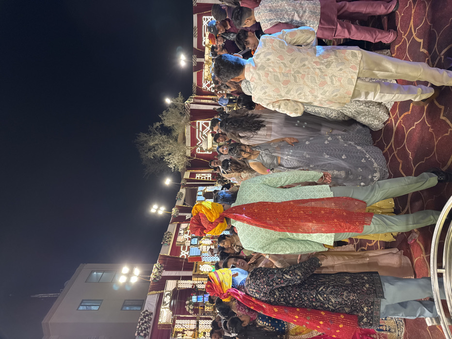 A nighttime outdoor gathering depicts guests in formal Indian attire. Men wear turbans and patterned sherwanis, while women are in embellished lehengas. Decorated arches frame the group, illuminated by numerous lights. The background showcases a building and a clear night sky. A patterned carpet covers the ground.