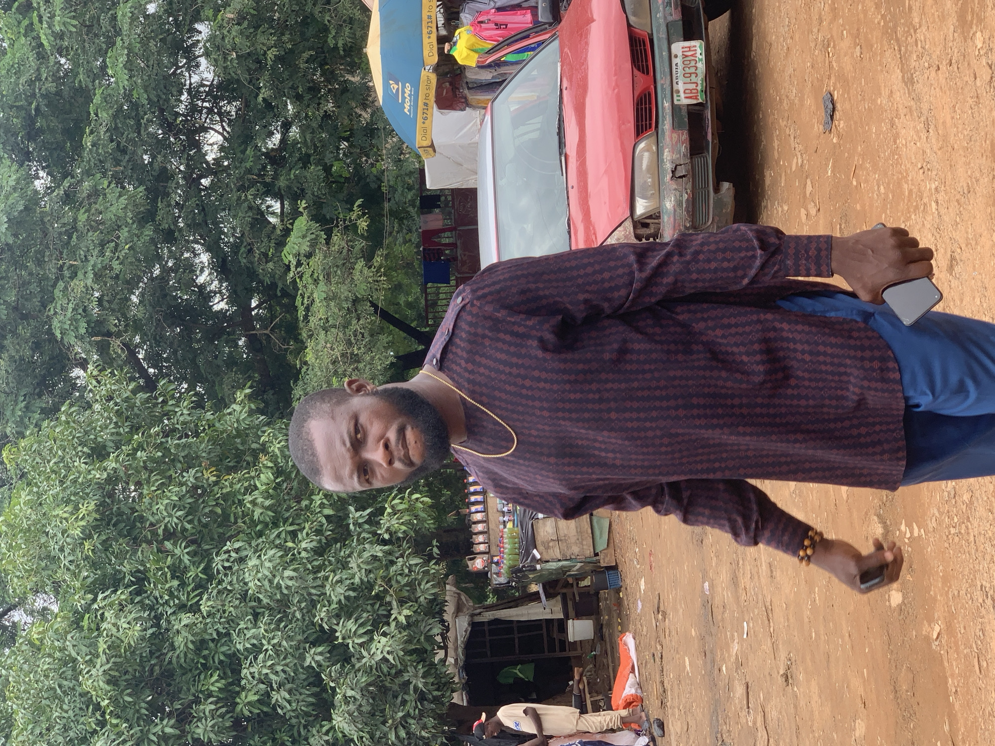 A man stands in an outdoor market, looking directly at the camera. He has dark skin, a beard, and is wearing a maroon and black patterned long-sleeved shirt with a gold chain and blue trousers. He is holding a dark-colored smartphone in his right hand. In the background, there are trees, makeshift stalls selling goods such as bottles of liquid and clothing. A red car with a Nigerian license plate that reads "ABJ-939XH" is parked behind him. The ground is dirt. The scene appears to be a typical street market in Abuja, Nigeria.