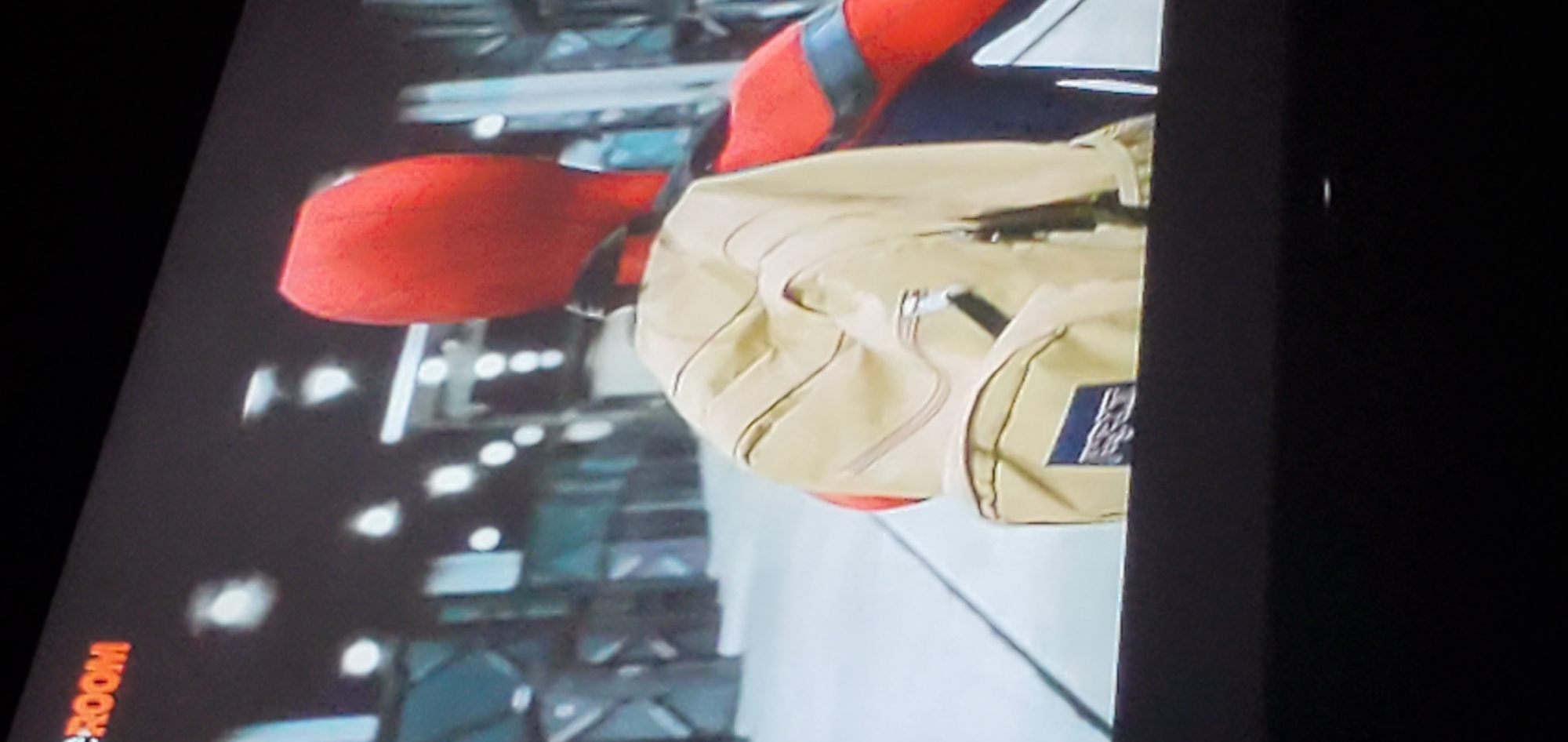 The image shows a person wearing a red suit, resembling Spider-Man, with a tan backpack. The person is seen from behind, facing away from the camera. The backpack has a blue label with white text that appears to be a brand logo. The background is blurry and shows what looks like an indoor environment with bright lights and structural elements. The top left corner of the image contains text that reads "CROOM" in orange. There is no information about the location in the city of Gura Top, Nigeria, nor any details that would suggest it.