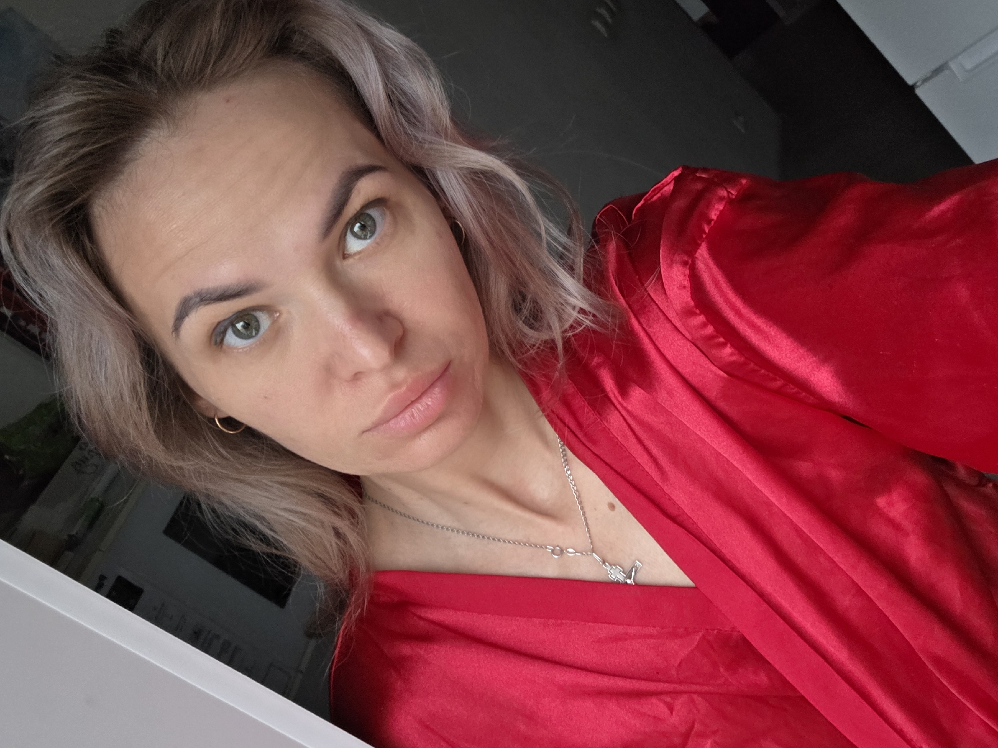 A woman is shown from a high angle in a close-up shot. She has light-colored hair with brown roots and fair skin. Her eyes are light green. She is wearing a red satin robe or shirt. Around her neck, she wears a silver necklace with a pendant. She has a gold hoop earring in one ear. The background is blurry, with some areas of black and white visible. Based on the provided information, the location is Chaikovsky, Russia.