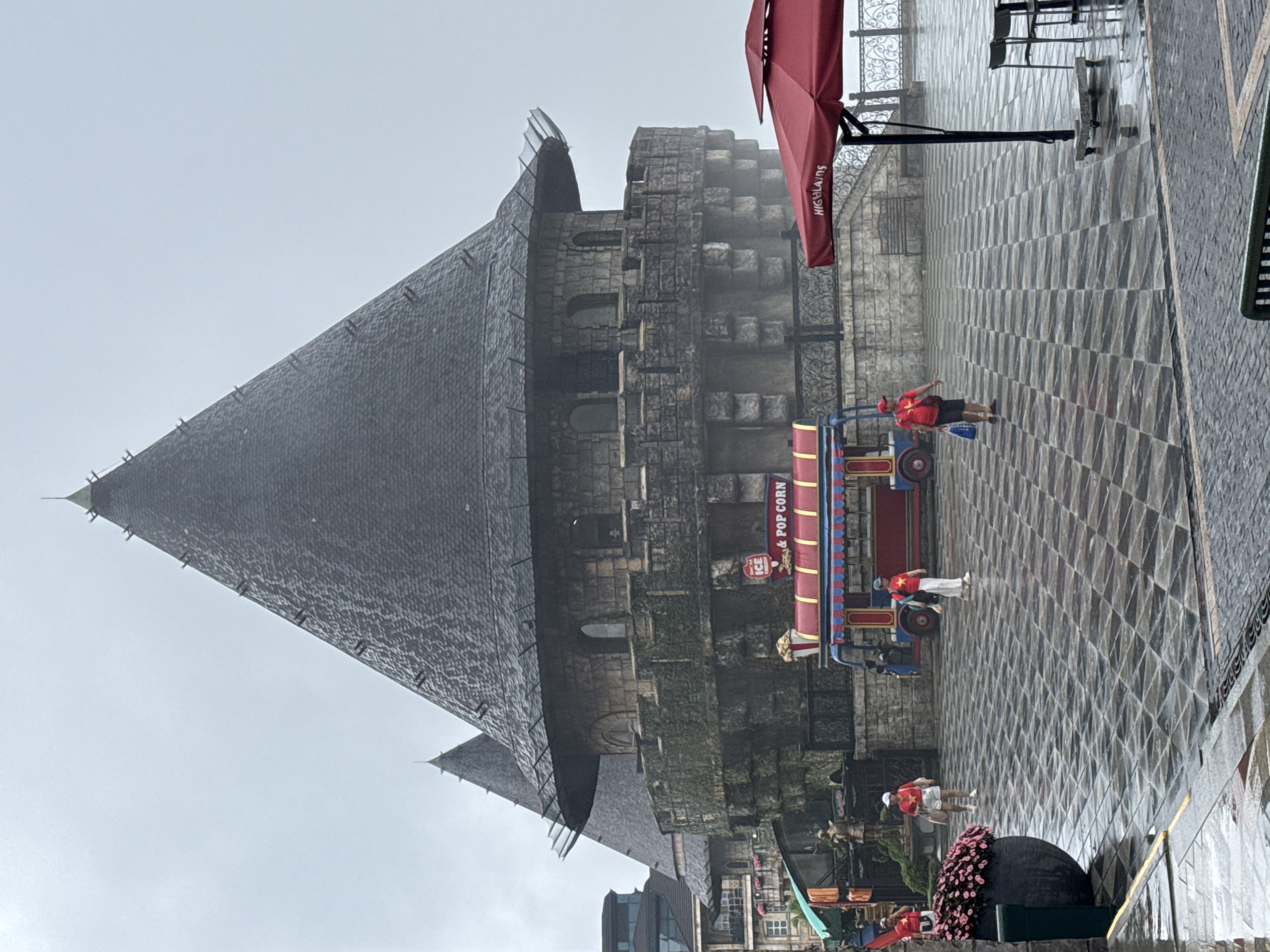 The image captures a rainy scene in Đà Nẵng, Vietnam, featuring the architecture of Ba Na Hills. The primary structure is a tall, gothic-style building with a steeply pitched roof covered in grey tiles. A portion of a red umbrella is visible in the upper-right corner. A small mobile cart selling ice cream and popcorn is positioned near the building. Several people wearing matching red shirts with yellow stars are moving around the area. The ground is covered in a pattern of grey paving stones. The weather appears overcast, with fog partially obscuring the upper parts of the building.