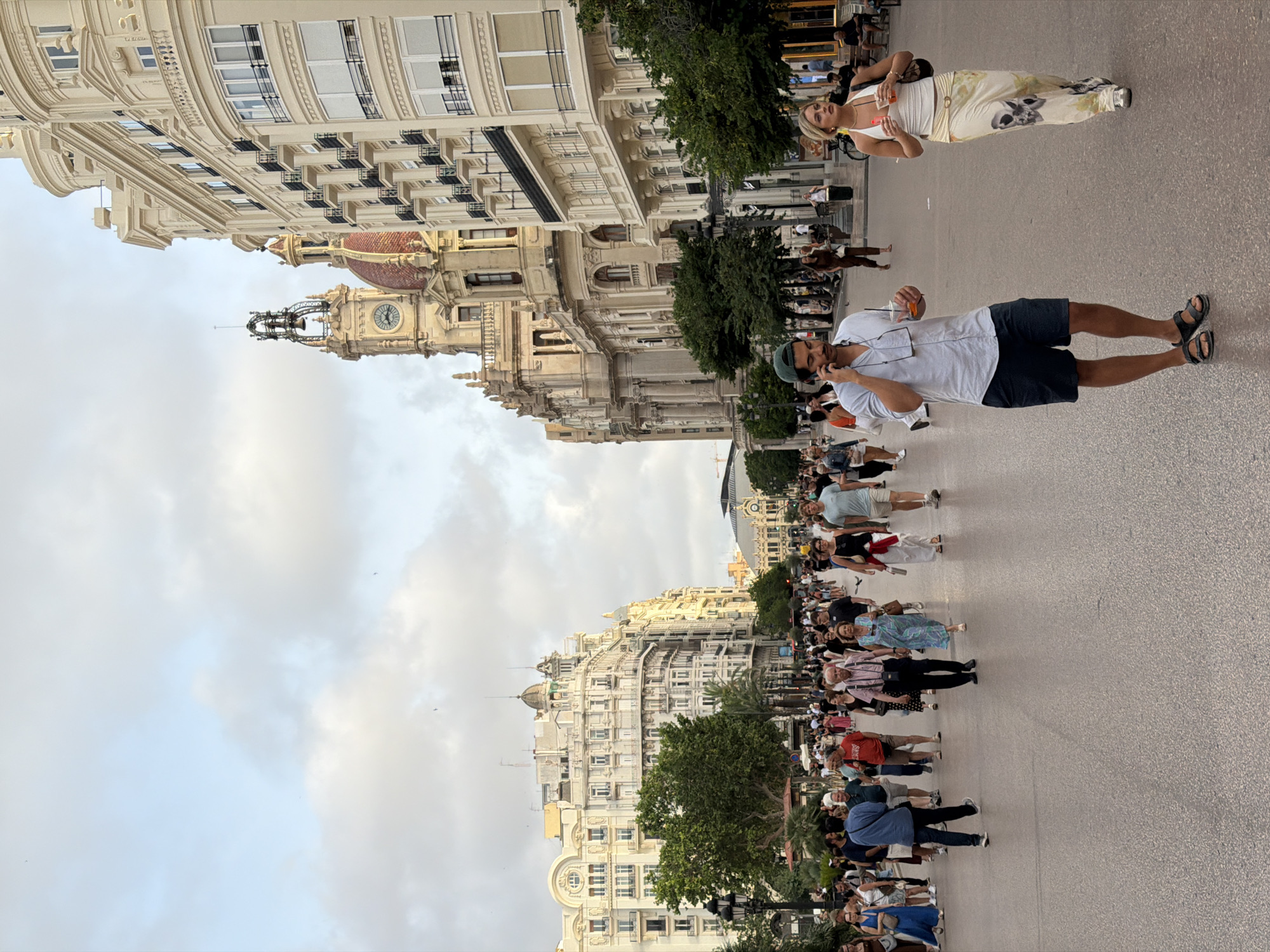 The image depicts a street scene in an urban environment, characterized by a wide pedestrian walkway lined with ornate, multi-story buildings. The architecture suggests a European city, possibly with historical significance, given the classical detailing on the buildings, including balconies, arched windows, and decorative cornices. One prominent building features a clock tower with a bell.
A diverse crowd of people is walking along the pedestrian area, engaged in various activities. Some are walking in groups, while others are alone. A man in the foreground, wearing a light blue shirt and dark shorts, is speaking on his phone and holding a drink. To his right, a woman in a white halter top and patterned pants is walking, also holding a drink. Other individuals in the background are dressed casually, suggesting a warm climate and a leisurely atmosphere.
The walkway is paved with a textured surface, and there are trees and lampposts interspersed along the sides, providing shade and lighting. The sky above is partly cloudy, with a soft light indicating it might be late afternoon or early evening. The overall impression is one of a bustling city square or promenade, likely a popular spot for locals and tourists.