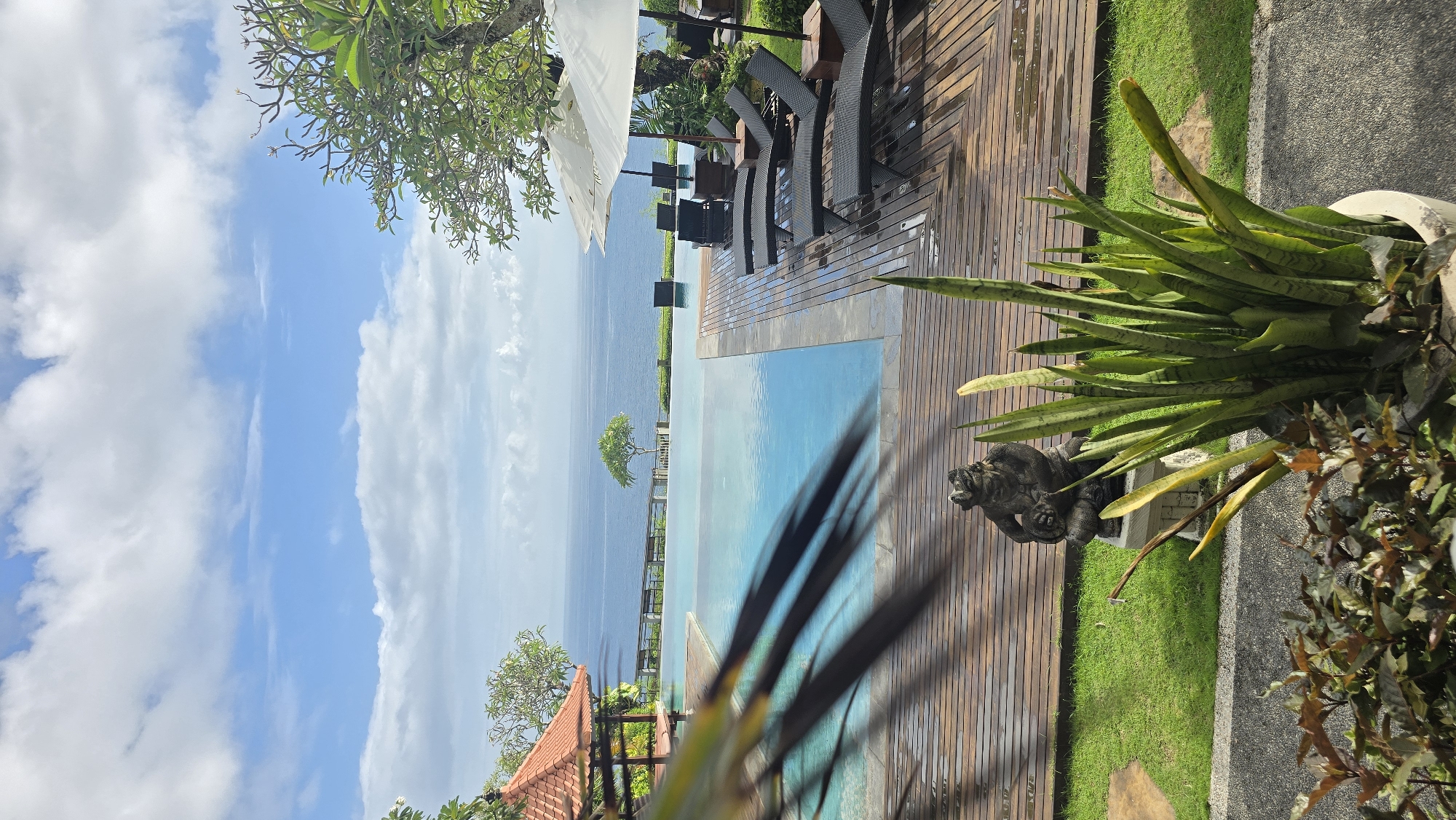 The image captures a tropical scene in Kuta Selatan, Indonesia, featuring an infinity pool overlooking the ocean. The pool is surrounded by a wooden deck with lounge chairs and umbrellas. In the foreground, there is lush vegetation, including snake plants and decorative statues. The sky is blue with scattered clouds. A building with a red tile roof is visible on the lower left. A small, tree-covered island is seen in the distance.