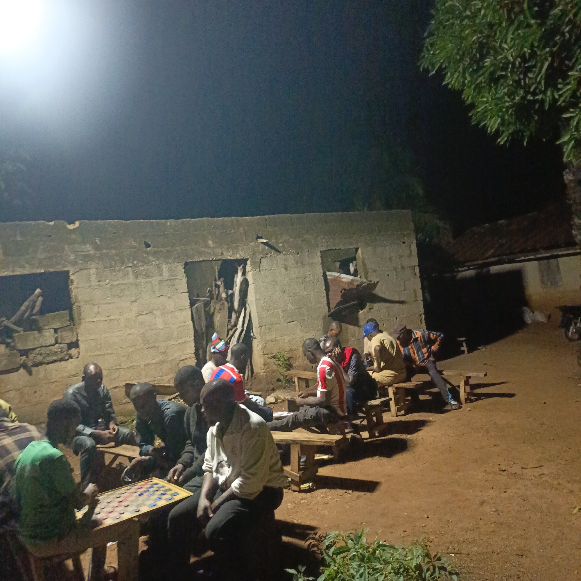 In Akwanga, Nigeria, at night, a group of men are gathered around a checkered board game. The scene is illuminated by an artificial light source casting a bright glow from the upper left corner of the frame, creating stark shadows. The men, dressed in casual clothing, are seated on wooden benches and stools, focused on the game laid out on a table in the foreground. The environment appears to be an outdoor space adjacent to a rustic, unfinished brick building. The ground is made of dirt and gravel, with some sparse vegetation visible. The overall atmosphere suggests a casual social gathering or pastime in a rural or semi-urban setting.