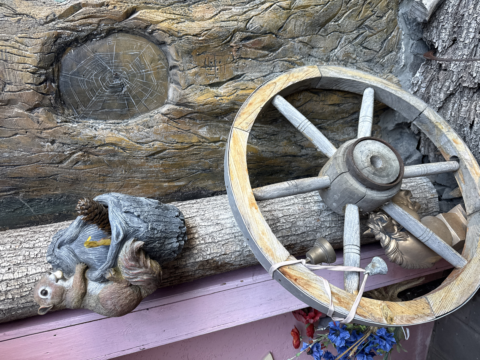 The image depicts a decorative arrangement of rustic elements against a textured, wood-like backdrop. In the upper left, a squirrel figurine is perched on a tree trunk, holding a small gray planter that contains a pine cone and what appears to be a yellow candle. To the right, the textured backdrop resembles tree bark with an inset oval shape displaying a white, web-like pattern. In the foreground, a weathered wooden wagon wheel is positioned diagonally, with a thin pink ribbon tied around its circumference. Two small brass bells and a charm are suspended from the ribbon. Beneath the wheel, a bronze-colored bust of a horse's head is partially visible, resting on a pedestal. A bouquet of artificial blue and red flowers is situated to the left of the wagon wheel. The environment appears to be an outdoor setting, possibly a garden or a decorative display. The location is identified as Okhtyrka, Ukraine.