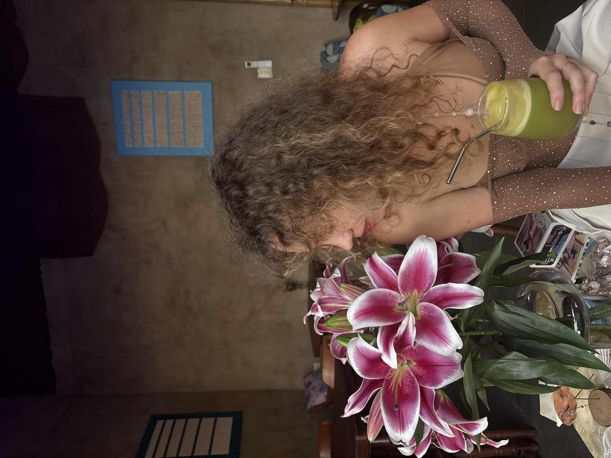 In Mũi Né, Vietnam, a woman with curly blonde hair is seated at a table in what appears to be a cafe or restaurant. She is wearing a tan top and sheer, jeweled sleeves, and is holding a glass of green juice with a metal straw. In front of her is a vase of pink and white lilies. The background shows tables, chairs, and decorative items on the wall, suggesting an indoor setting with a rustic or bohemian ambiance.