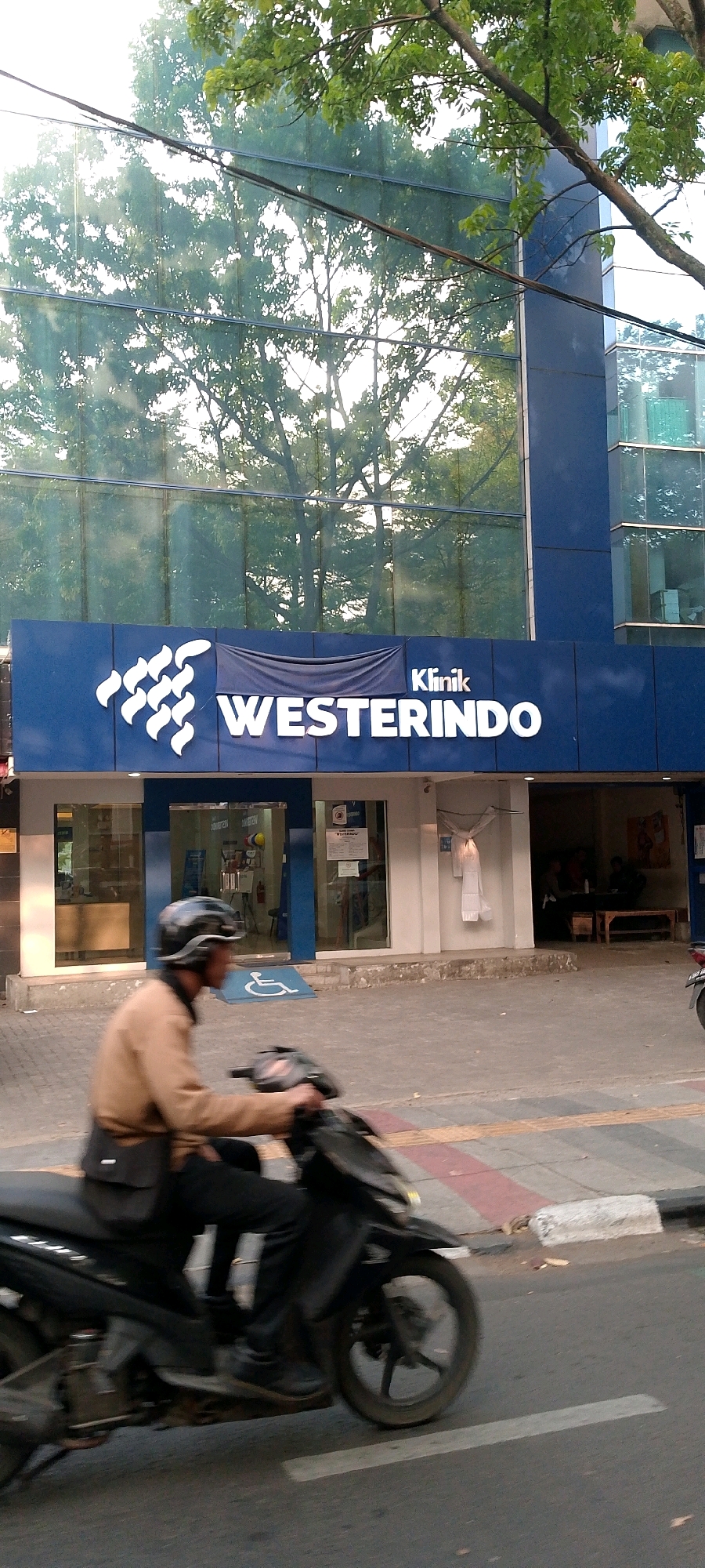 The image shows the facade of a clinic named "WESTERINDO" with the word "Klinik" above it. The building has a blue exterior with large glass windows reflecting trees and the sky. A person is riding a black motorcycle past the building. The motorcycle is in motion, and the rider is wearing a tan jacket and a helmet. A blue ramp with a white wheelchair symbol is visible on the sidewalk in front of the clinic entrance. The street in front of the clinic has pedestrian walkways with red and white striping. The background shows more of the building and trees. Based on visual cues from the surrounding architecture and the presence of a clinic named "WESTERINDO", this location is likely in Bandung City, Indonesia.