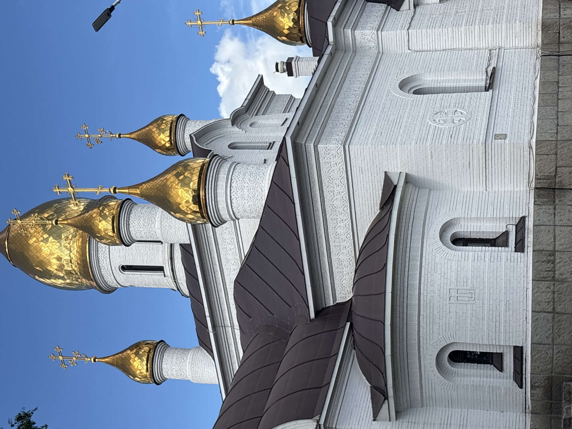 This media file depicts a close-up, angled view of the exterior of a white brick church with golden domes. The church is a prominent subject, with its ornate architecture and shimmering domes dominating the frame. Several golden domes are visible, each topped with a cross. The white brick walls are textured and feature decorative carvings and arched windows. Dark brown, segmented roofs are visible between the domes and the main body of the church. The sky is a clear, bright blue, with a few white clouds present, suggesting fair weather. A street lamp or light fixture is visible in the upper portion of the frame, silhouetted against the sky.
The scene is likely set during the daytime, given the bright lighting and blue sky. The architecture strongly suggests a Russian Orthodox church. While there are no people or overt activities depicted, the image captures the grandeur and religious significance of the building. The focus is on the structural and decorative elements of the church itself, set against a serene sky. No text is visible in the image.