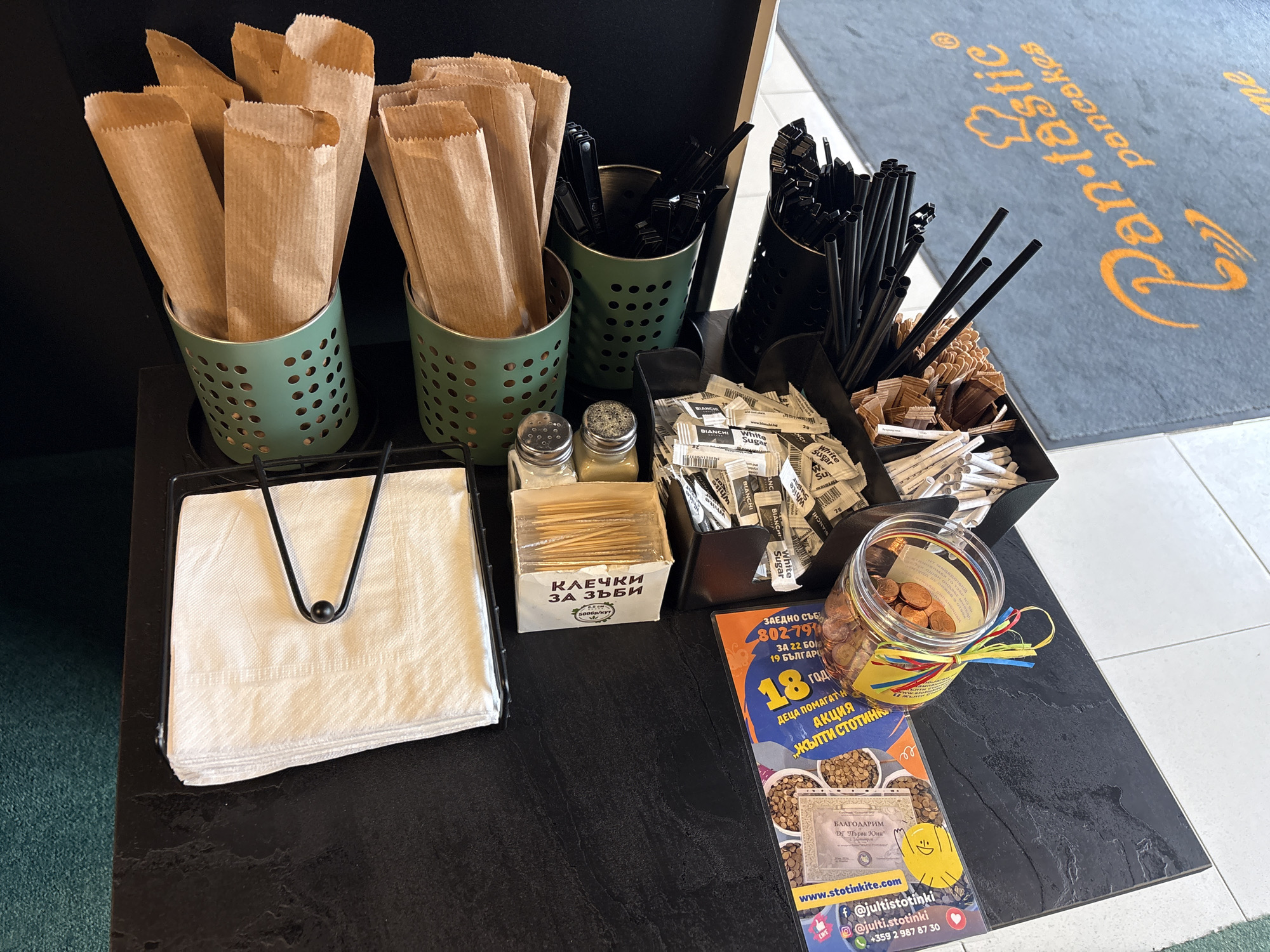 The image depicts a tabletop arrangement in Veliko Tarnovo, Bulgaria, likely within or near the "Pan-tastic pancakes" establishment based on the logo on the floor mat visible in the upper right corner. The scene is a service station for customers. There are several green, perforated metal containers holding brown paper utensil sleeves. Additionally, there is a black container holding various black straws and stir sticks, next to smaller containers holding condiments such as sugar and toothpick boxes labeled "КЛЕЧКИ ЗА ЗЪБИ". A napkin holder with white napkins is also present. A glass jar filled with coins, decorated with ribbons, and a flyer displaying donation efforts by "ФГ "Първи Юни"", including the website "www.stotinkite.com" with social media handles and a Bulgarian phone number are visible. The tabletop itself has a dark, textured surface.