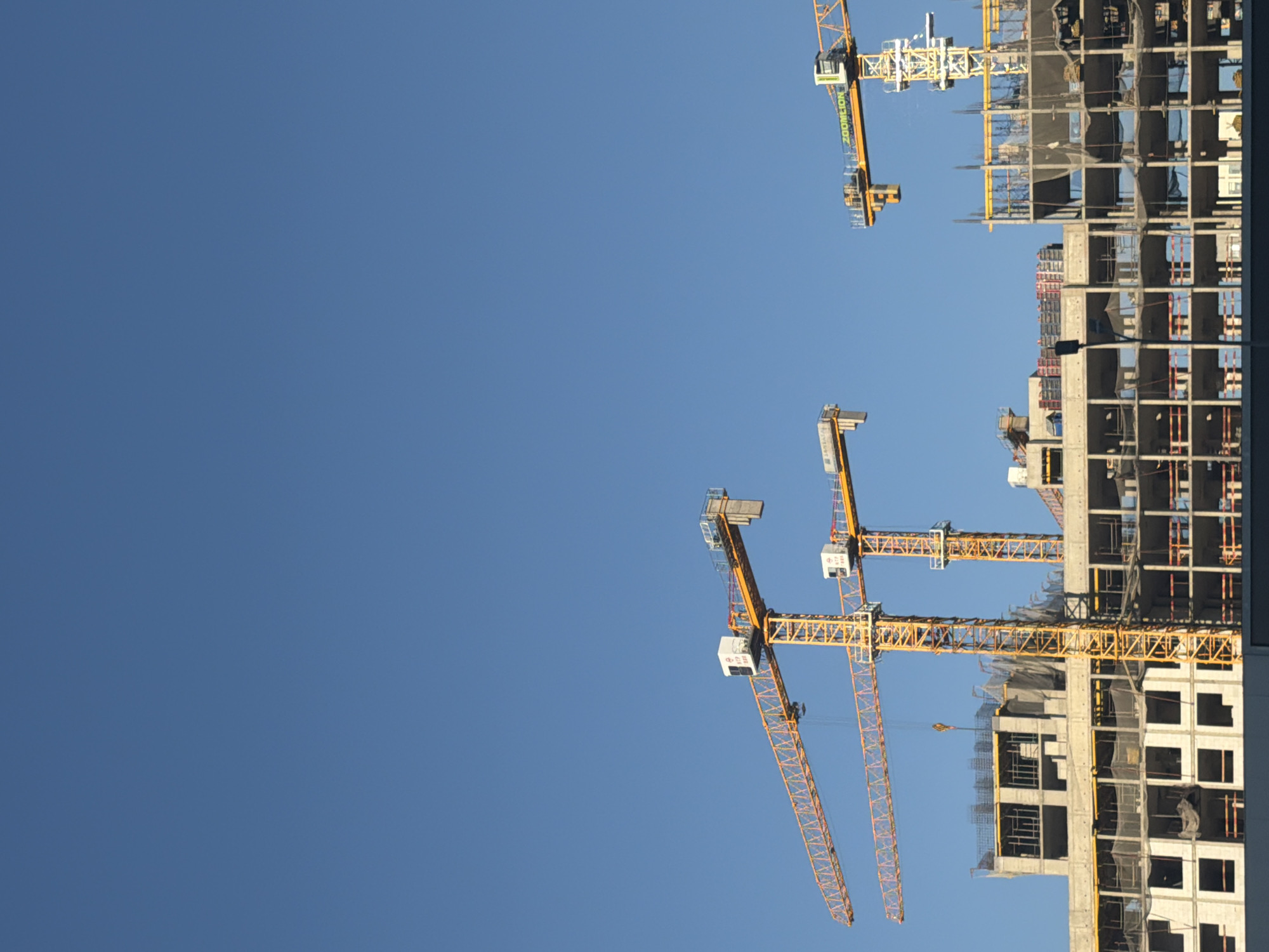 This is a photograph of a construction site with multiple cranes against a clear blue sky. The construction site features several buildings in various stages of completion, with exposed concrete structures and scaffolding. Three large yellow tower cranes are prominently visible, positioned over and around the buildings. One crane is in the foreground on the left, another is in the mid-ground, and a third is to the right. The cranes appear to be in operational use, as indicated by their configuration and the presence of construction activity on the buildings. The overall scene suggests ongoing urban development. The image appears to be taken in Astana, Kazakhstan, as indicated by an inscription on one of the cranes that reads "ZOOMILION," a brand of construction equipment commonly used in the region and associated with construction projects in Astana.