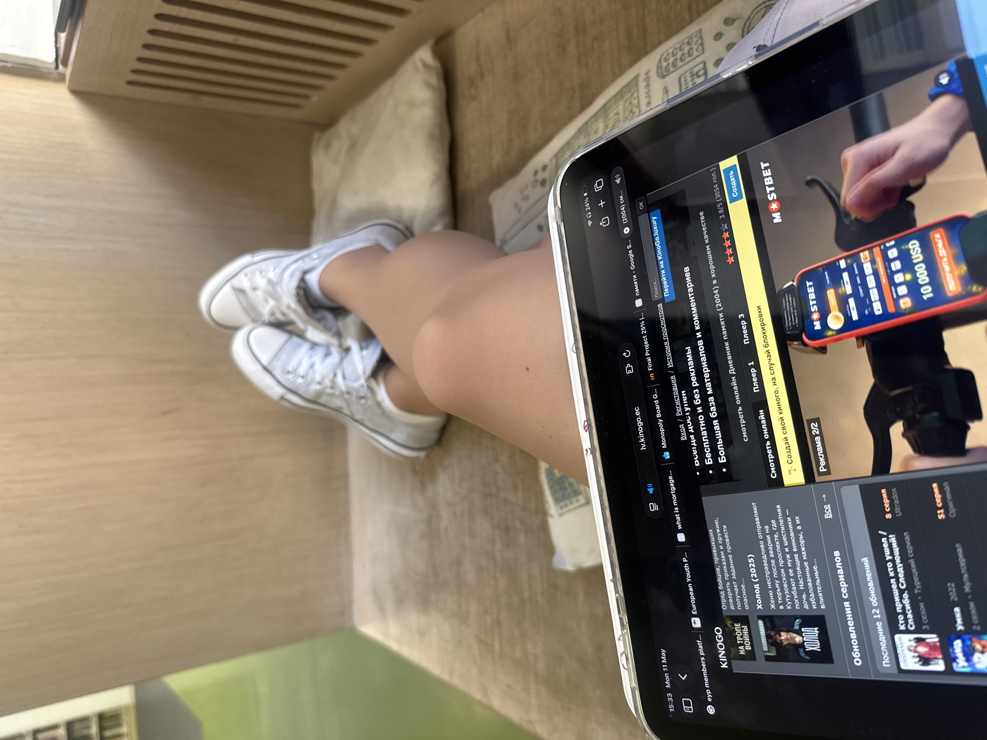 The image shows a person sitting indoors, likely on a cushioned bench or sofa. They are wearing white sneakers and appear to be using a tablet. The tablet screen displays what looks like a streaming service or website with movie/series options. There is an ad banner at the top for MostBet and another ad showing a hand holding a phone with a 10,000 USD notification. The time displayed on the screen is 15:33, and the date is Mon 11 May. It is impossible to confirm the location in Yerevan, Armenia without additional clues.