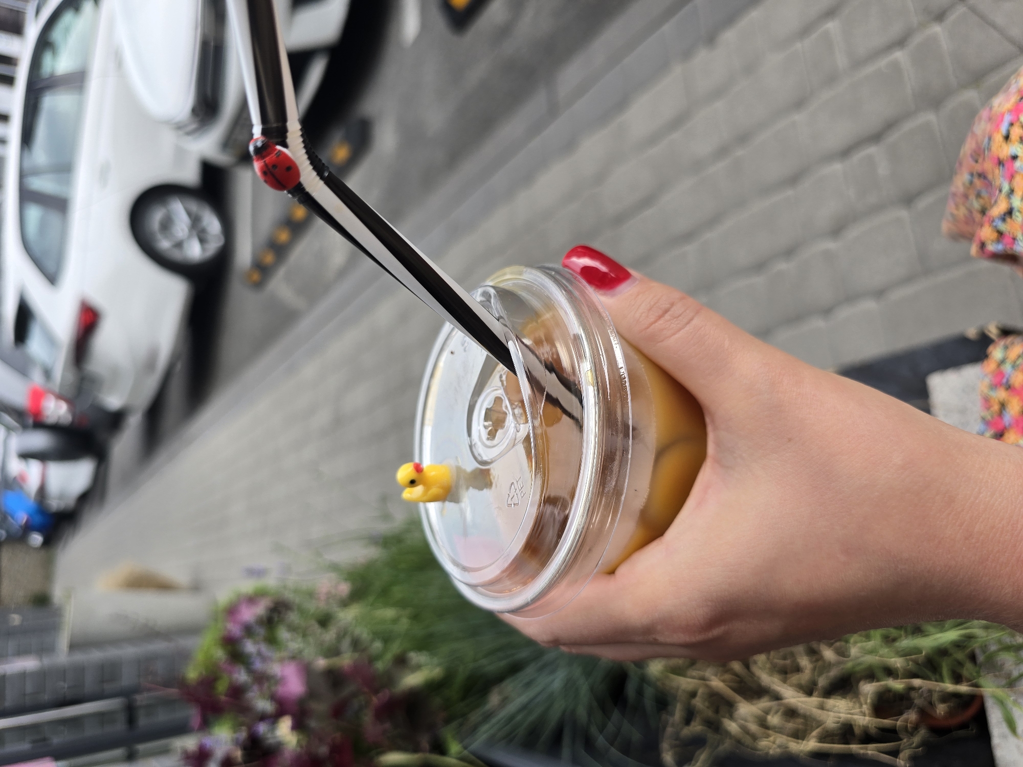 A person's hand with red-painted fingernails holds a clear plastic cup with a yellow drink and a lid. A black and white striped straw is inserted into the lid, decorated with a red ladybug charm and a small yellow duck figure. The cup is positioned over a blurry background of green plants and a grey brick wall. In the upper left, a blurry white car is visible. The image appears to have been taken outdoors during daylight. The lighting suggests it might be a sunny day. The setting seems to be urban or suburban, possibly a cafe or a street scene. The overall impression is casual, with a focus on the beverage and its playful decorations. The hand and cup are in sharp focus, while the background elements are intentionally blurred, drawing attention to the drink.