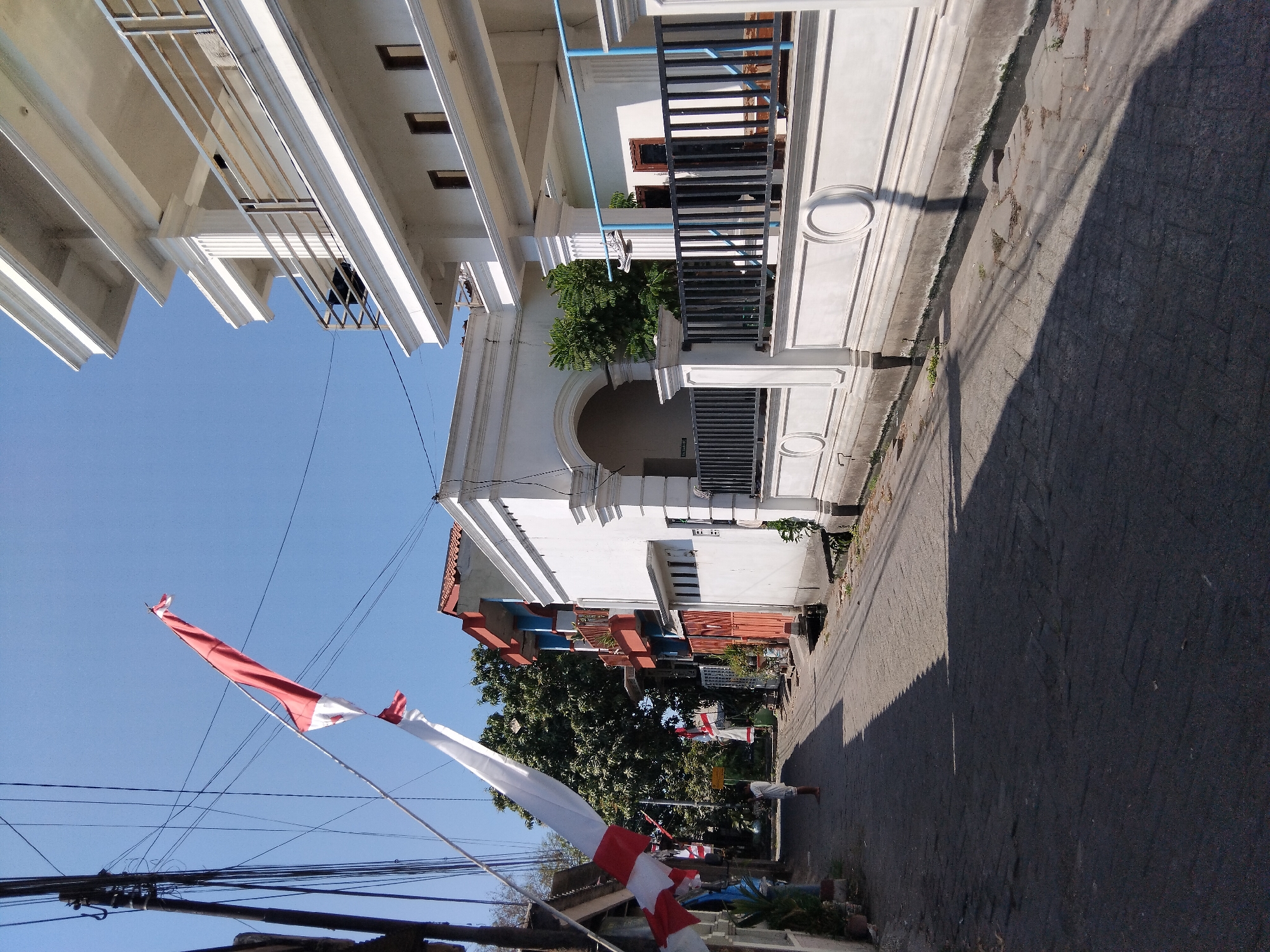 In the city of Berbek, Indonesia, a narrow, paved street is depicted on a sunny day. Red and white Indonesian flags are strung across the street, fluttering in the breeze. On the right side of the street, a modern, multi-story white building with balconies and a black metal fence is visible. To the left of the street, several other buildings are present, including a white one with an arched entrance and a multi-story structure with red and blue accents. The street is lined with trees and various plants. A lone figure, wearing white clothing, walks away from the camera down the street. The ground is paved with interlocking stones and is partially covered by the shadows cast by the buildings and flags. Electrical wires are strung overhead, supported by poles.