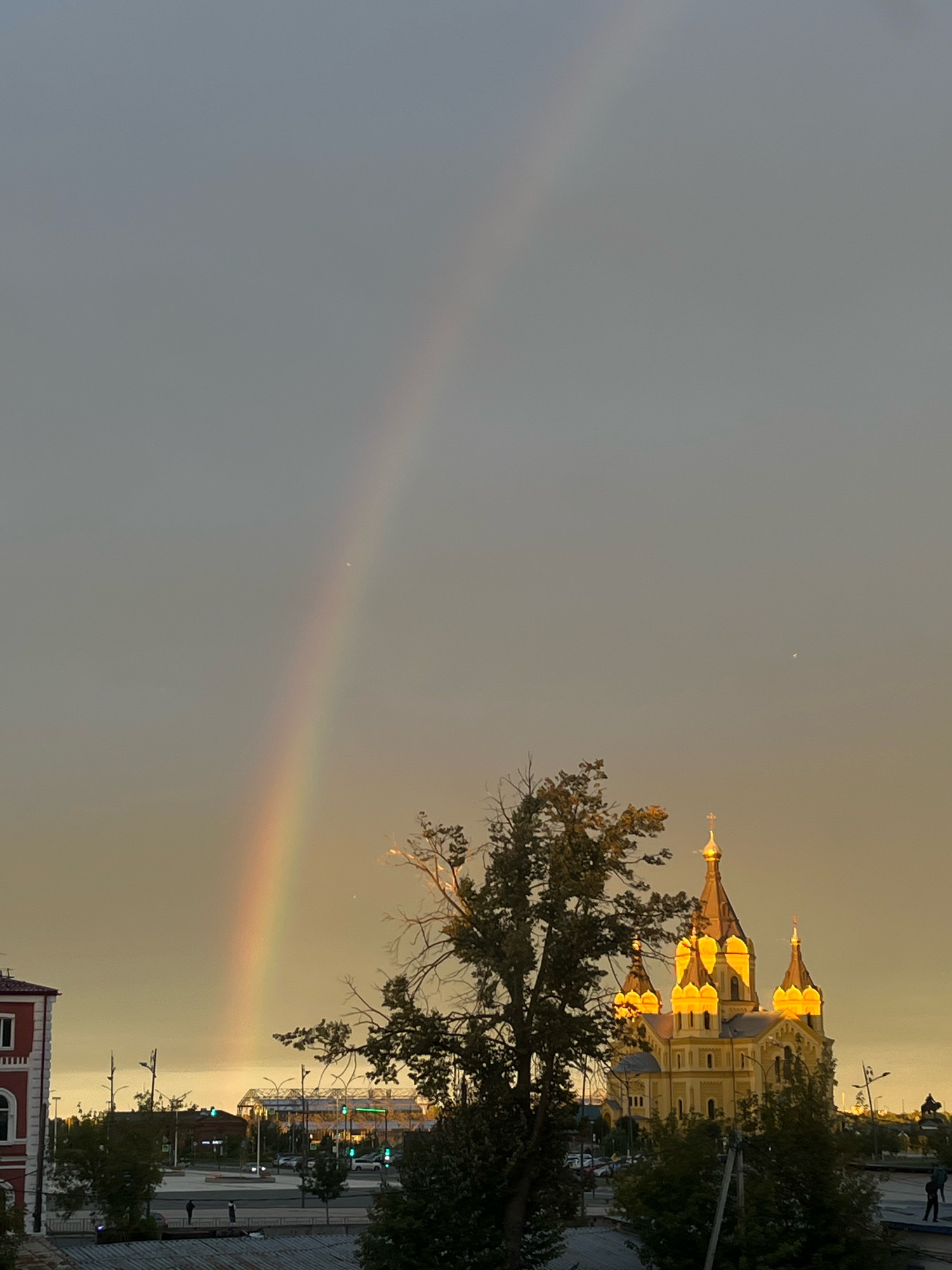 The image captures a view of the Alexander Nevsky Cathedral in Nizhny Novgorod, Russia, under a sky displaying a rainbow and faint celestial bodies. The cathedral, illuminated by warm, artificial lighting that highlights its golden domes and architectural details, stands prominently in the mid-ground. Surrounding the cathedral are trees with dense foliage, partially obscuring the structure. In the foreground, a tree with sparser branches is visible on the right side. To the left, a red building with white-trimmed windows is partially visible. The background shows a city landscape with streetlights, traffic lights (some green), vehicles, and a few pedestrians in the distance. The sky is overcast with a muted gray tone, except for the bright arc of the rainbow stretching across it.