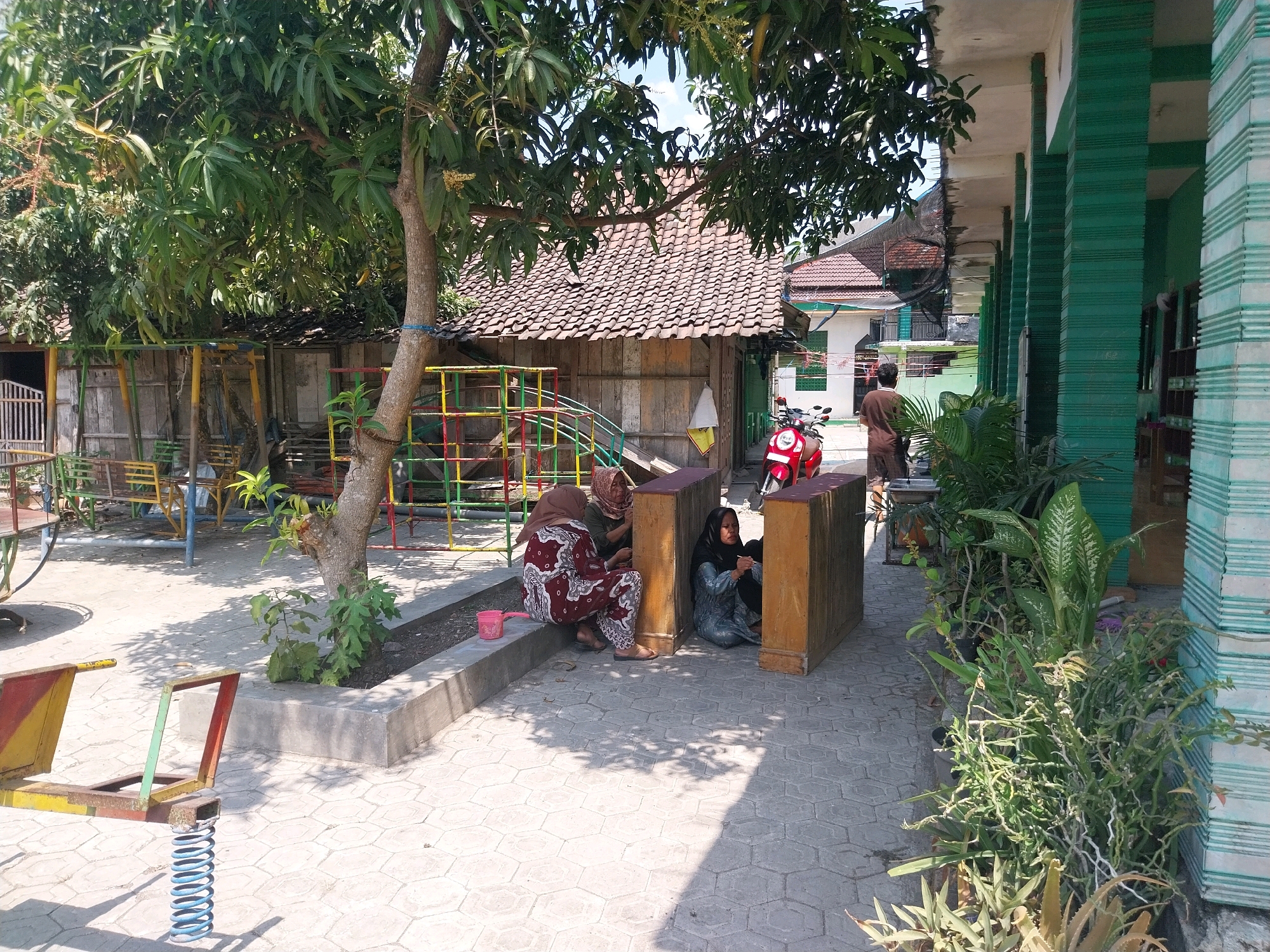 The image depicts an outdoor scene on a sunny day, likely in a schoolyard or community area in Campur, Indonesia. In the foreground, a children's playground structure with a blue spring is visible, along with a paved walkway. To the left, a weathered wooden building with a tiled roof is situated, partially shaded by a large tree with leafy branches. Adjacent to this building is a playground area with colorful metal frames and swings.
In the middle ground, three women are seated on the ground near a wooden structure. Two of the women are wearing hijabs and traditional Indonesian attire. One woman in a patterned maroon and white outfit is seated on a raised concrete planter. Another woman in a green hijab and patterned outfit is seated next to her. A third woman, wearing a black hijab and a blue patterned dress, is seated to the right, leaning against the wooden structure. A red scooter is parked behind them.
To the right of the image, a building with green striped pillars and a white roof extends along a walkway. Lush green plants and potted vegetation are arranged along the walkway. In the background, more buildings with tiled roofs are visible, suggesting a residential or institutional setting within a town or village. The overall impression is of a community space with elements of both recreation and daily life.