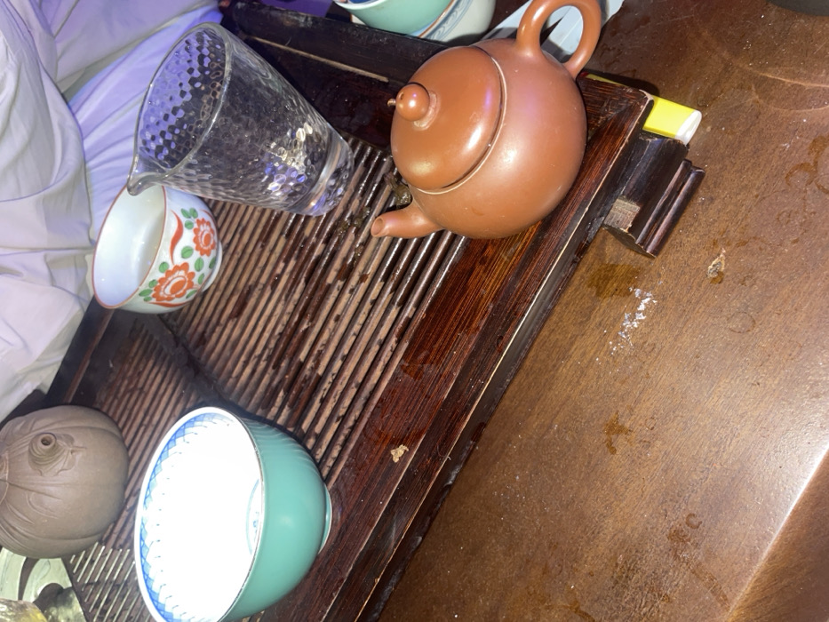 The image displays a close-up, elevated view of a tea ceremony setup, presumably in Nha Trang, Vietnam. No individuals are present. The primary subjects are various ceramic and glass vessels arranged on a dark brown wooden tea tray with a slatted surface, designed to drain excess liquid. The tray and the darker wooden table it rests upon are wet with water spills and show white residue, indicative of recent use.
From left to right on the tea tray:
1. A large, teal-colored ceramic bowl with a white interior and a blue geometric pattern along its rim. It appears empty.
2. Behind the teal bowl, a light brown ceramic object shaped like a gourd or pumpkin, with a small spout, is partially visible.
3. A smaller white ceramic bowl with an orange floral pattern, containing a small amount of clear liquid.
4. A clear, textured glass pitcher or decanter, upright, showing reflections of light.
5. A small, reddish-brown earthenware teapot with a lid and spout is positioned towards the lower right of the tray.
The background consists of blurred white fabric, possibly upholstery or bedding, illuminated with a distinct purple hue, suggesting artificial lighting conditions in an indoor setting. The overall scene indicates the recent preparation or consumption of tea.