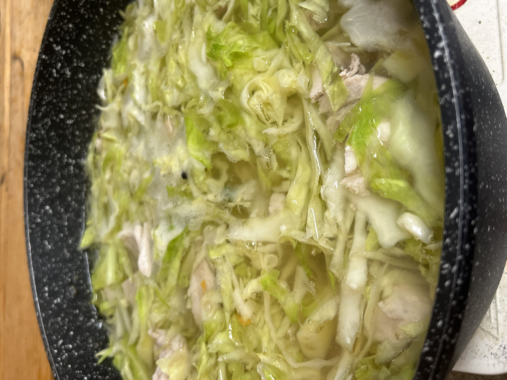 A close-up, overhead shot shows a black pot filled with a light-colored broth containing shredded cabbage and pieces of meat. The broth is bubbling, indicating it is hot and likely cooking. The cabbage is bright green and appears tender, while the meat pieces are white and pinkish. Some smaller, lighter-colored ingredients, possibly potatoes or daikon radish, are also visible. The pot has a textured, speckled black exterior. The background shows a light-colored wooden surface, likely a countertop or table. No people or city landmarks are visible, so no specific location in Vasilyevo, Russia can be determined.