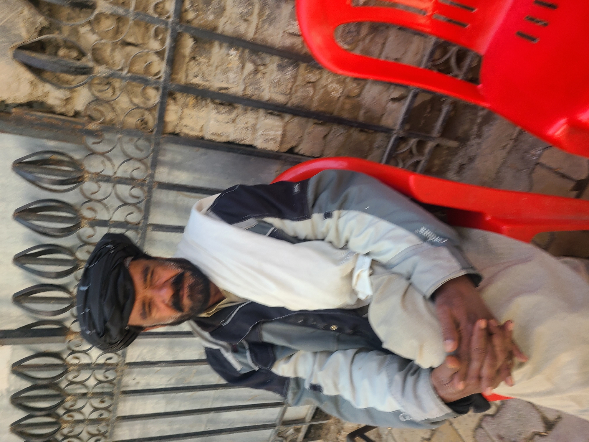 A man is sitting in a red plastic chair outside, likely in Quetta City Tehsil, Pakistan. He wears a black turban and a jacket with light gray and black sections, and a white wrap. His hands are clasped together. The man is next to a wrought iron fence. The setting appears to be urban, with a brick wall visible in the background.