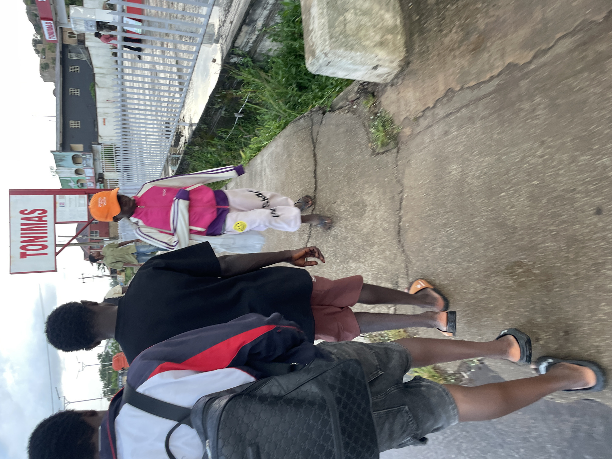 The image depicts a street scene in Jos, Nigeria. In the foreground, three young individuals, likely male, are walking away from the camera on a wet, cracked concrete walkway. The person in the center wears a black t-shirt and red shorts. To their left, another individual wears a black backpack, a white and red jacket, and dark shorts. To the right of the central figure, a fourth person, appearing to be a younger individual, is wearing a bright pink and purple jacket, white pants with smiley face emblems, and an orange baseball cap. The background shows a row of buildings, one with a prominent "TONIMAS" sign. A white metal fence separates the walkway from a more built-up area. The sky is overcast. The overall impression is of a candid street snapshot taken during daylight hours, possibly after rain.