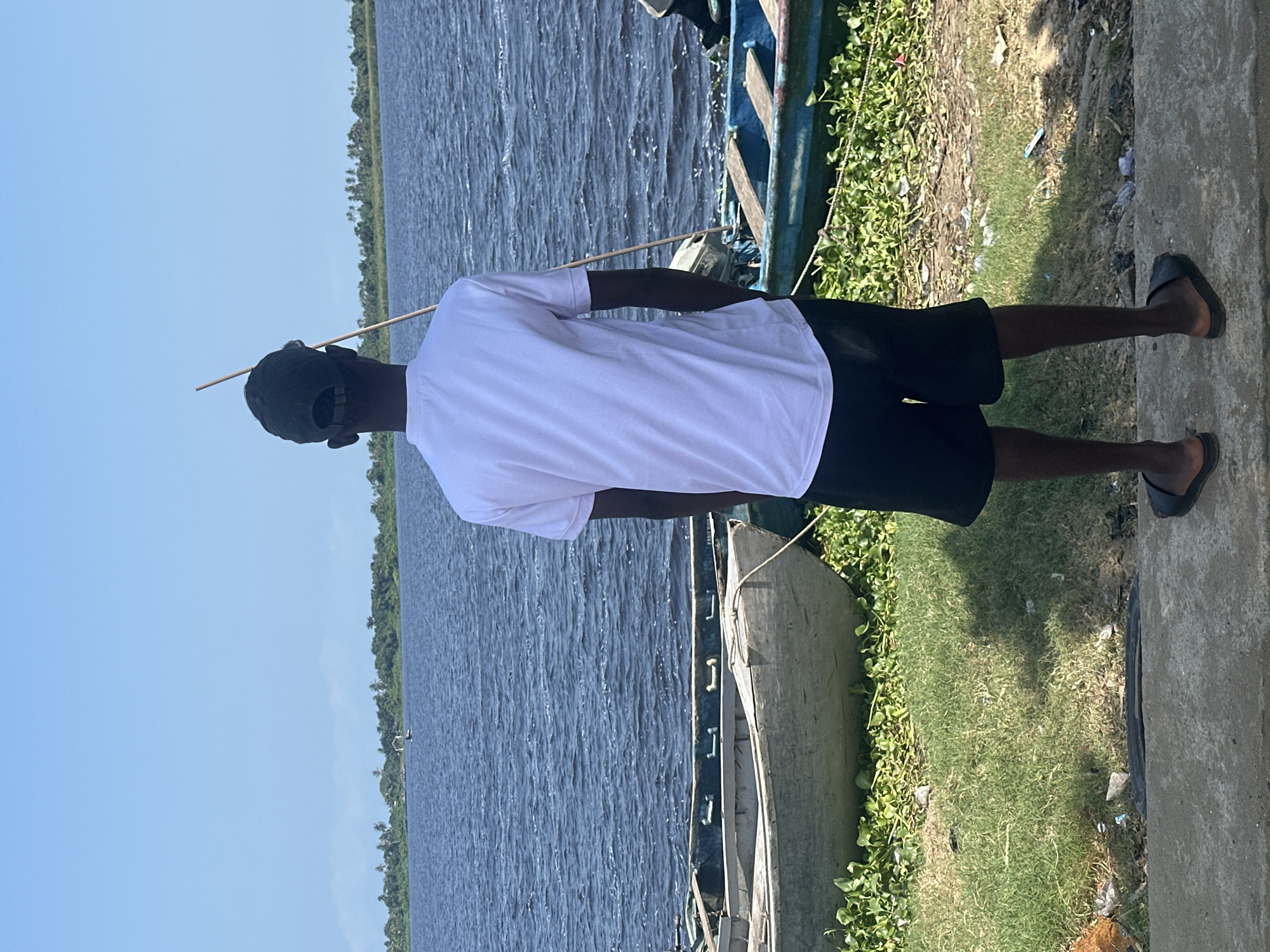 In Badagry, Nigeria, a young Black male stands on a concrete embankment, facing a wide, rippling body of water under a clear blue sky. He is wearing a white t-shirt, black shorts, a dark baseball cap, and black slides. He holds a thin, light-colored pole in his right hand, angled upwards and slightly behind him. In the foreground, several weathered wooden boats are moored on the grassy bank, some partially submerged in aquatic vegetation. The water extends to a distant tree-lined horizon. The scene captures a moment of quiet contemplation by the waterfront.