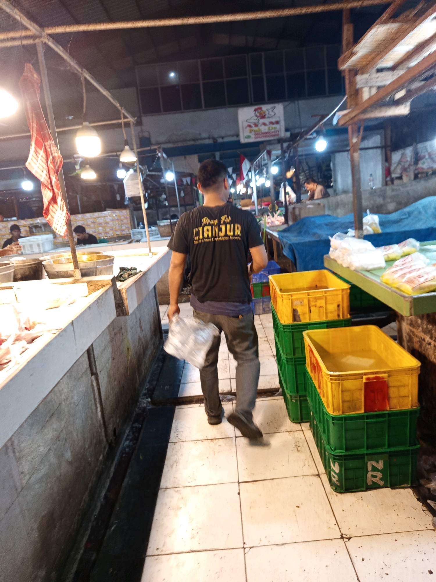 The image captures a bustling scene inside a night market, likely in Karawang, Indonesia, given the context provided. A man, seen from behind, walks away from the camera, holding a plastic bag. He is wearing a black t-shirt with text that appears to read "DRAMA" and "CIANJUR" in yellow and orange lettering. His pants are a muted grey, and he is wearing dark shoes. The market environment is characterized by overhead fluorescent lighting illuminating various stalls and goods. To the right of the man, a stack of green and yellow plastic crates is visible, some containing items. In the background, other individuals can be seen tending to their stalls, and market wares are displayed, though details are obscured by the dim lighting and distance. The floor is tiled, showing signs of wear and occasional dirt. The overall atmosphere suggests a lively commercial space, active during the evening hours.