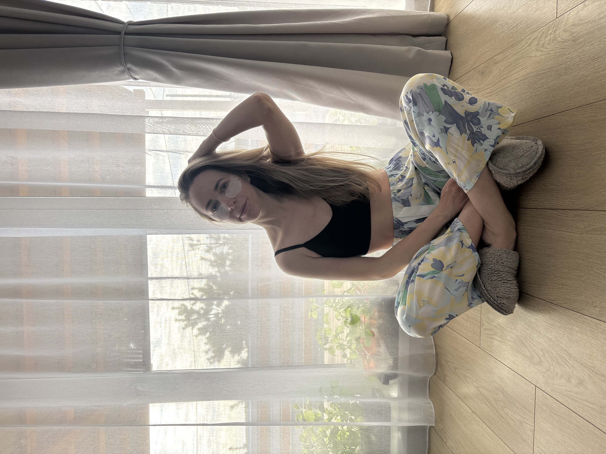 A woman is seated on a light brown wooden floor in front of a window. She is wearing a black crop top, patterned pajama pants, and gray slippers. She has blonde hair and is wearing eye patches. Her left hand is raised, touching her hair. The window is covered with sheer white curtains, partially open, revealing a glimpse of greenery outside. There are heavier gray curtains pulled back at the top of the window. The location is Chaikovsky, Russia.