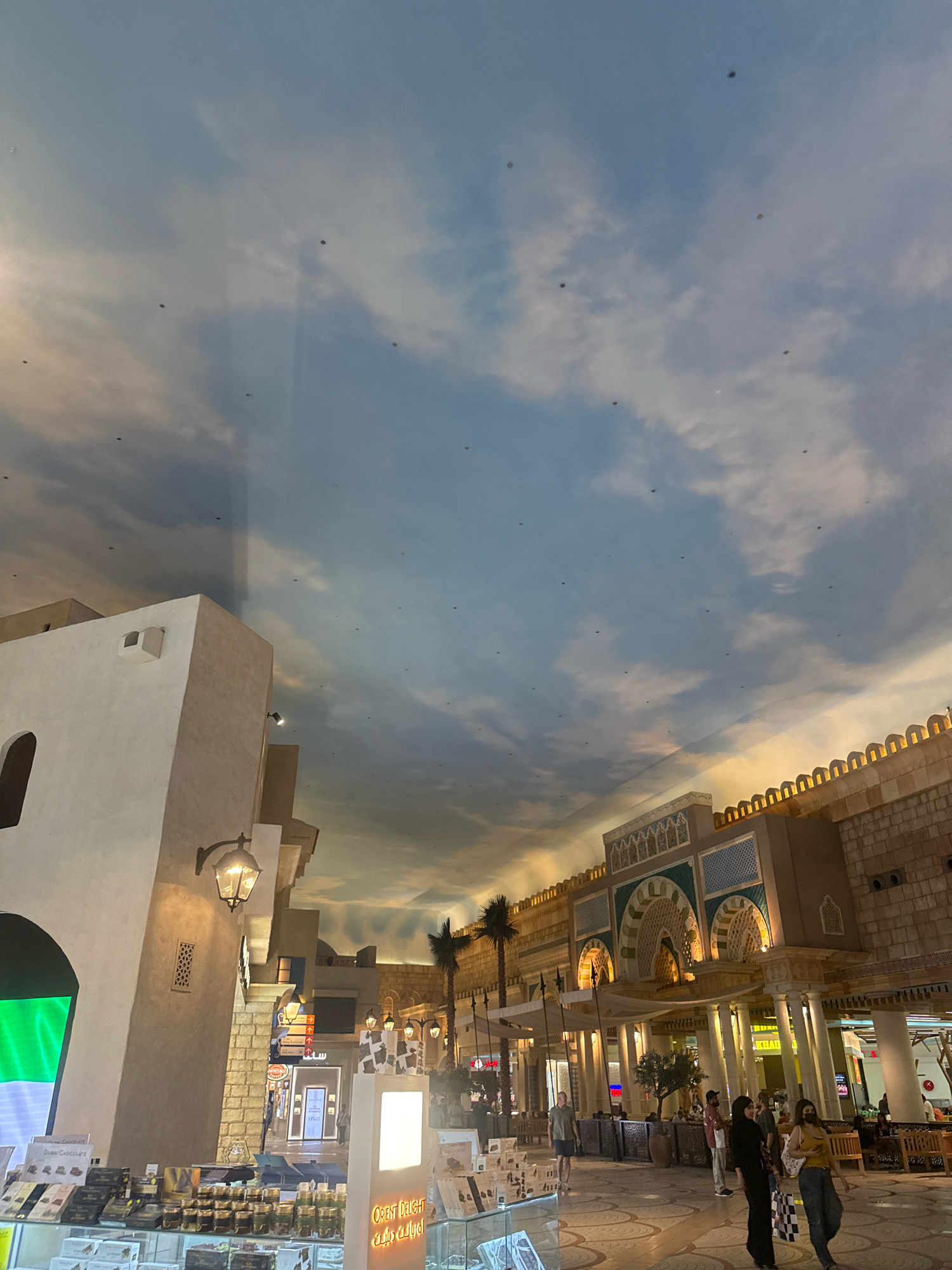 The image depicts an indoor shopping environment designed to resemble a traditional Middle Eastern marketplace, possibly within a Dubai mall such as the Mall of the Emirates or Ibn Battuta Mall, given the architectural style. The ceiling is a painted mural of a cloudy sky, giving the illusion of an outdoor setting. Several small, dark objects are scattered across the sky mural, which appear to be dust or debris.
In the foreground, to the left, is a storefront displaying numerous boxes and jars of what appears to be chocolates and sweets. The signage indicates "Dubai Chocolate" and "Orient Delight" in English and Arabic. A bright green and white fabric display is also visible to the left of the storefront.
Further into the scene, several people are walking through the marketplace. To the right, a building with a facade featuring ornate arches and decorative tile work, reminiscent of Islamic architecture, stands prominently. This structure is illuminated by warm lighting. Palm trees are strategically placed to enhance the market atmosphere. People are seen browsing shops and walking along the tiled pathway. The overall impression is a retail space designed for an immersive shopping experience.