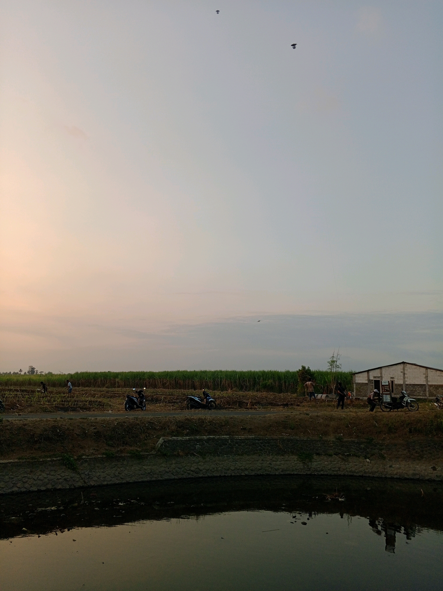 The image depicts an outdoor scene in what appears to be a rural area near Gondanglegi, Indonesia, during either dawn or dusk, given the soft, warm light. In the foreground, a dark, still body of water with reflections of the sky and surrounding objects occupies the lower portion of the frame. The water is bordered by a textured concrete embankment.
Above the embankment, a dirt road runs horizontally across the mid-ground. Along this road, there are several people and objects. Two motorcycles, one dark and one lighter colored, are parked on or near the road. A group of people, some appearing to be agricultural workers, are visible near a low-lying building constructed from white bricks. The building has a dark roof and an open doorway. Further back, a field of tall, green crops, possibly sugarcane, stretches across the landscape.
The sky above is a gradient of pale orange and light blue, with scattered clouds. Two dark, silhouette-like objects are visible high in the sky, possibly birds or kites. The overall impression is one of a quiet, everyday scene in a cultivated landscape.