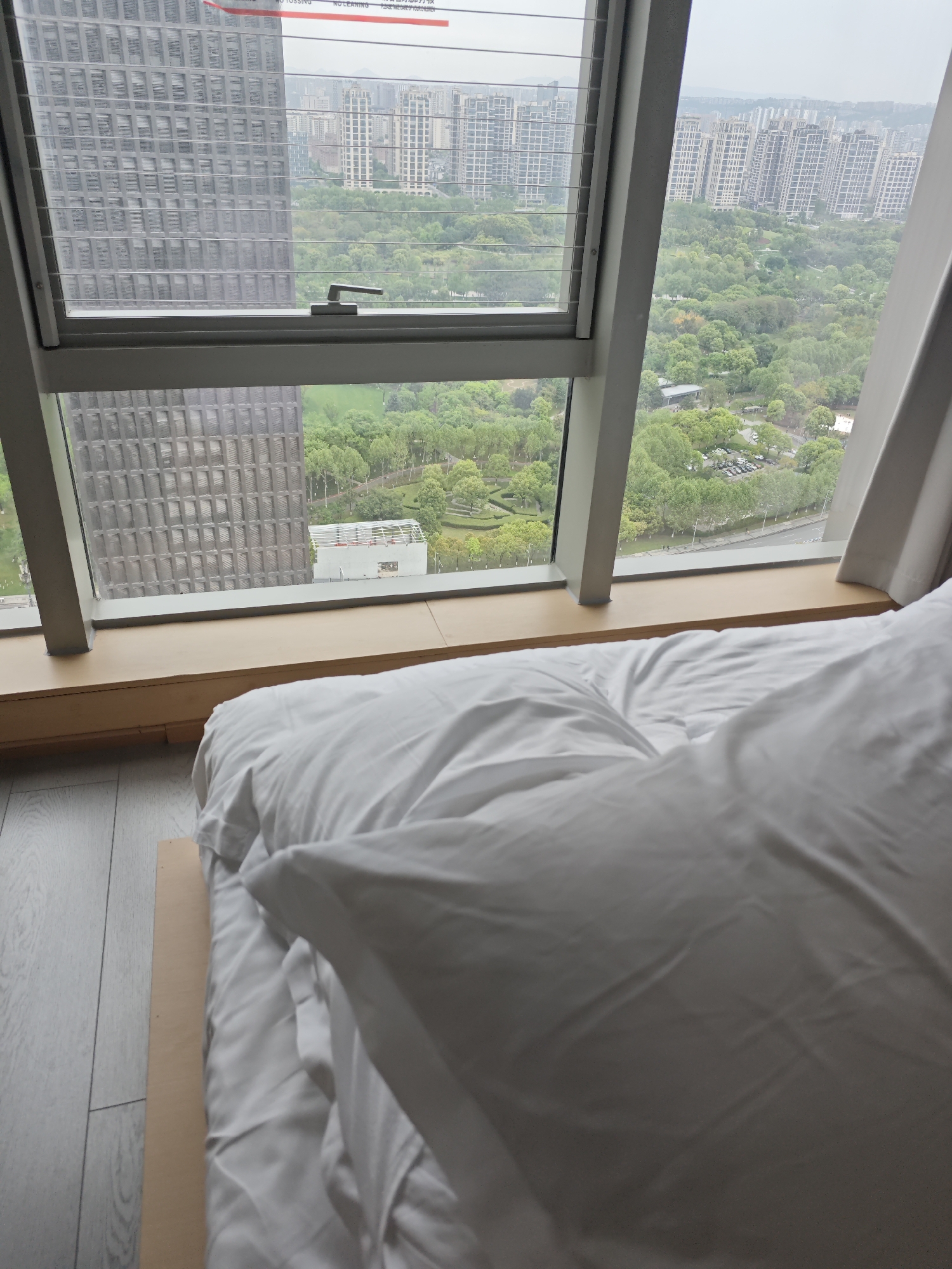 The image depicts a view from a high-rise window in what appears to be a hotel room. In the foreground, a bed with white linens and a large pillow takes up much of the scene. The window provides a view of an urban landscape, featuring other high-rise buildings in the distance and a park-like area with greenery and trees in the middle ground. The sky appears overcast or foggy.
Based on the architecture and overall urban landscape, the image is likely to be taken in Liangjiang, China. There are no people visible in the image. The primary objects are the bed, the window, and the urban landscape. The focus of the image is on the view outside the window.