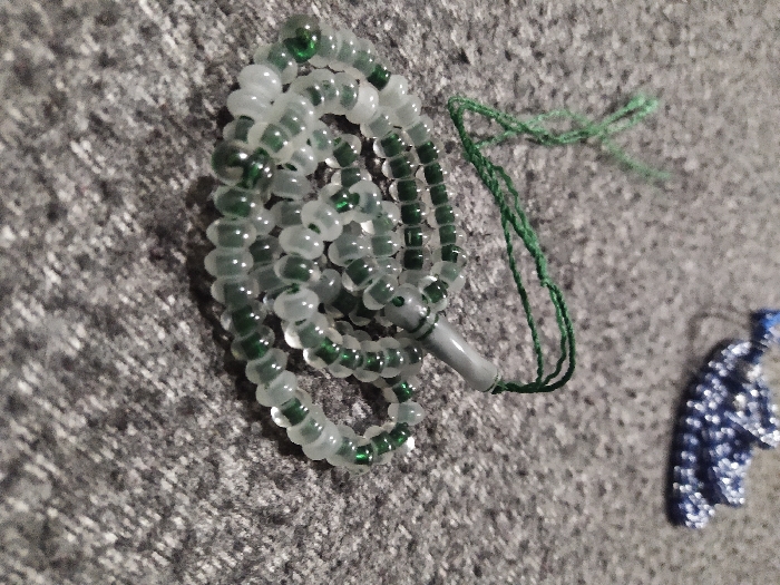 The scene displays a set of prayer beads loosely coiled on a mottled gray, textured surface, likely fabric, in the city of Unknown, Unknown. The primary subject consists of transparent beads alternating between clear/white and dark green. A distinct grayish-white cylindrical "imam" bead is present, from which a dark green multi-strand string or tassel extends. In the upper right background, a second, out-of-focus object, appearing to be another smaller, coiled item with blue and white coloration, is visible. No individuals are present, and no actions or interactions are depicted. The environment appears to be an interior setting.