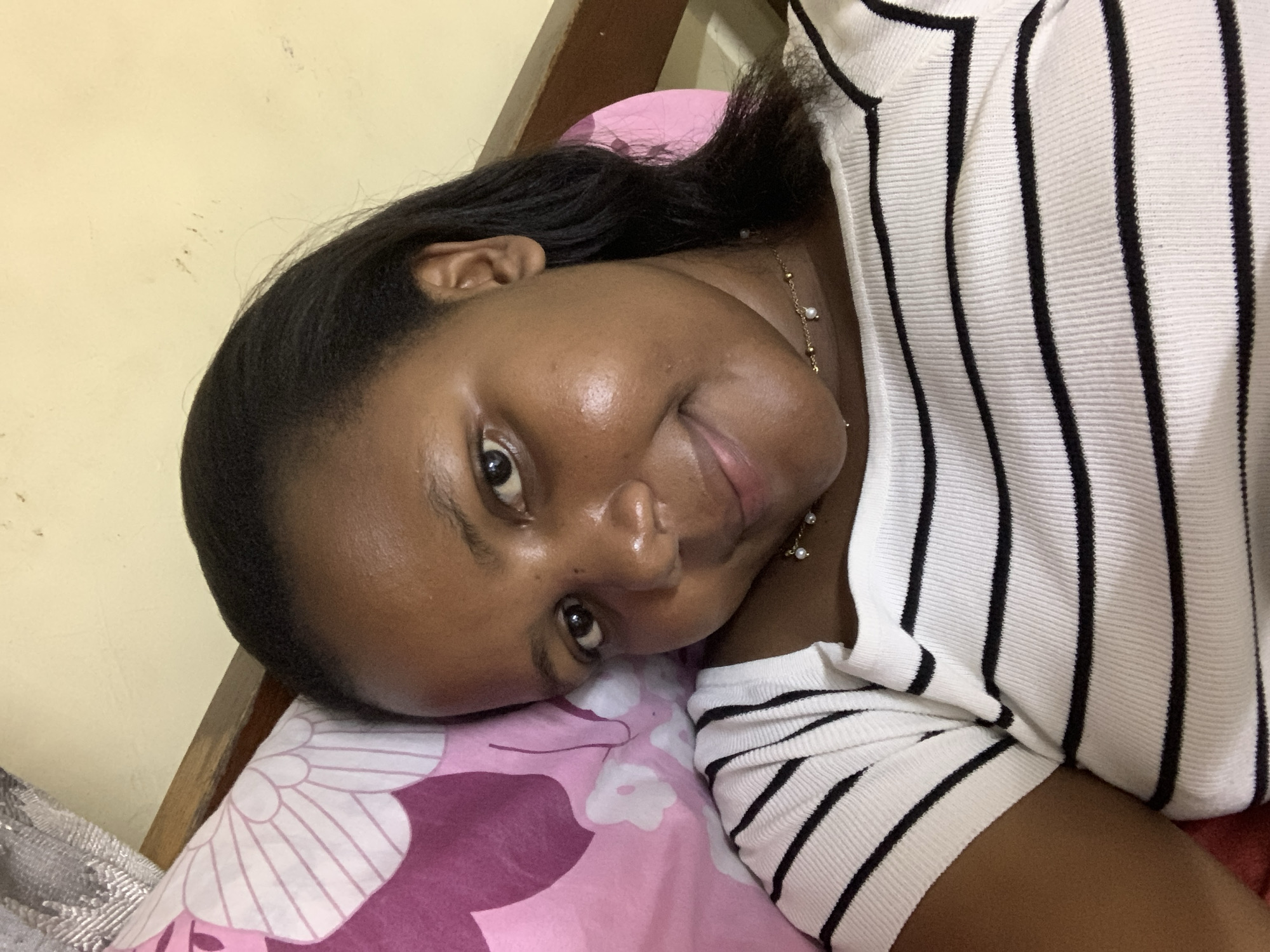 A dark-skinned woman with black hair is lying on a pink pillow in a bed. She is wearing a white shirt with black stripes and a gold necklace. The background is a yellow wall. The location cannot be determined from this image.