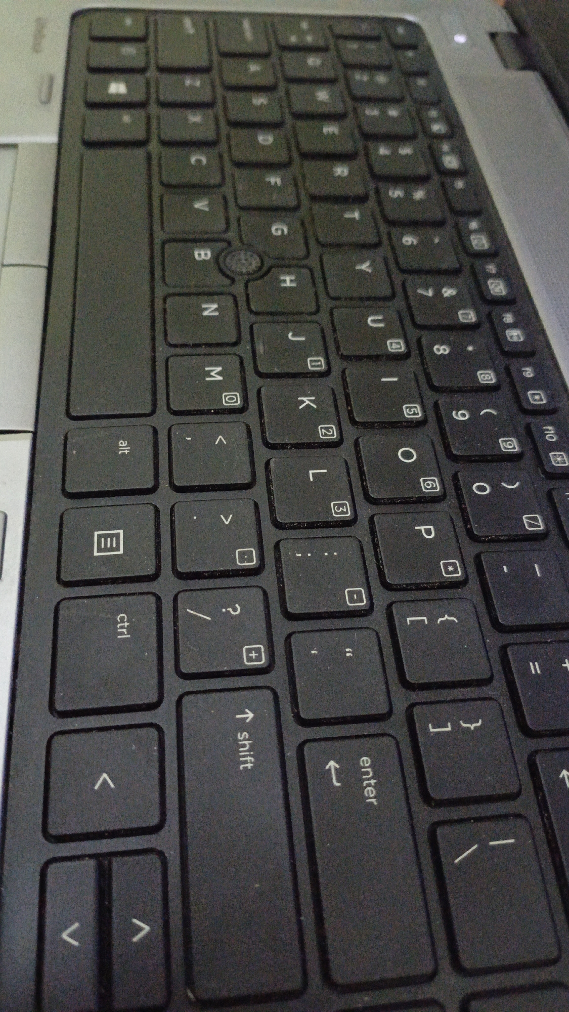 The image shows a close-up, angled view of a black laptop keyboard. The keys are black with white lettering. Some keys have symbols or smaller numbers above the primary letter. The laptop's silver casing is visible on the right side and top edge of the image. The keyboard is angled and the perspective is from above, looking down and slightly to the side. There are no people or any indication of the specific location in Trihanggo, Indonesia.