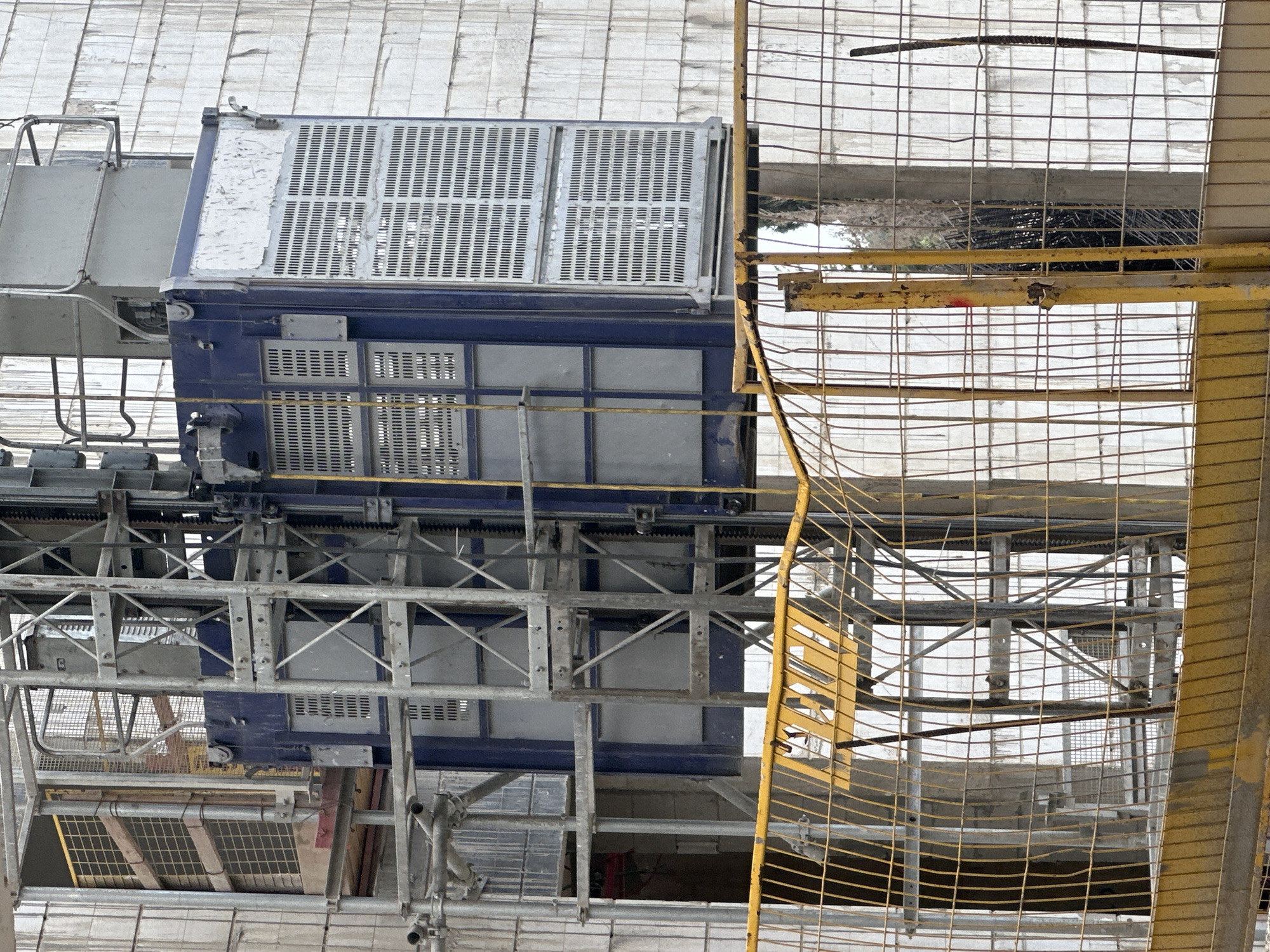An outdoor, medium-angle shot depicts a construction site in Haifa, Israel, focusing on a temporary elevator attached to a building under construction. The elevator, painted blue and silver, is a rectangular structure with grated sides, positioned vertically along a metal scaffolding system. It appears to be a hoist or personnel lift, commonly used in high-rise construction. The building's exterior is clad in light-colored, textured blocks. A yellow metal safety fence in the foreground partially obscures the view of the construction area. The scene is devoid of people, suggesting the photograph was taken during a non-operational period. The overall impression is one of ongoing construction work in an urban environment.